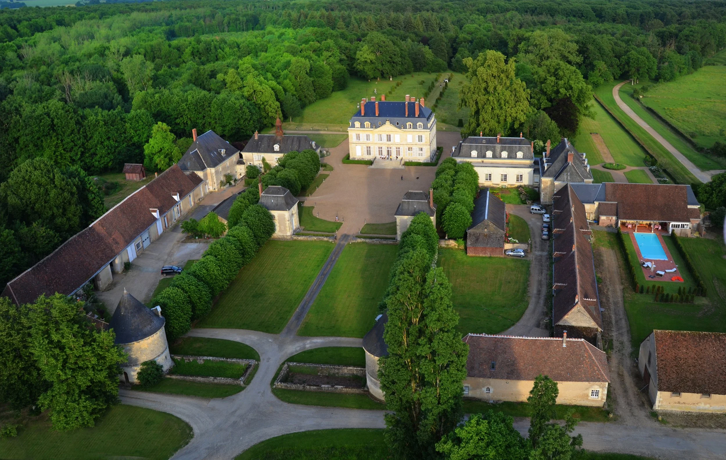 An aerial view of a large historic estate with a central grand building, surrounded by smaller buildings, lush trees, and well-maintained gardens and lawns.  Venue is privately held but can host exclusive luxury events.