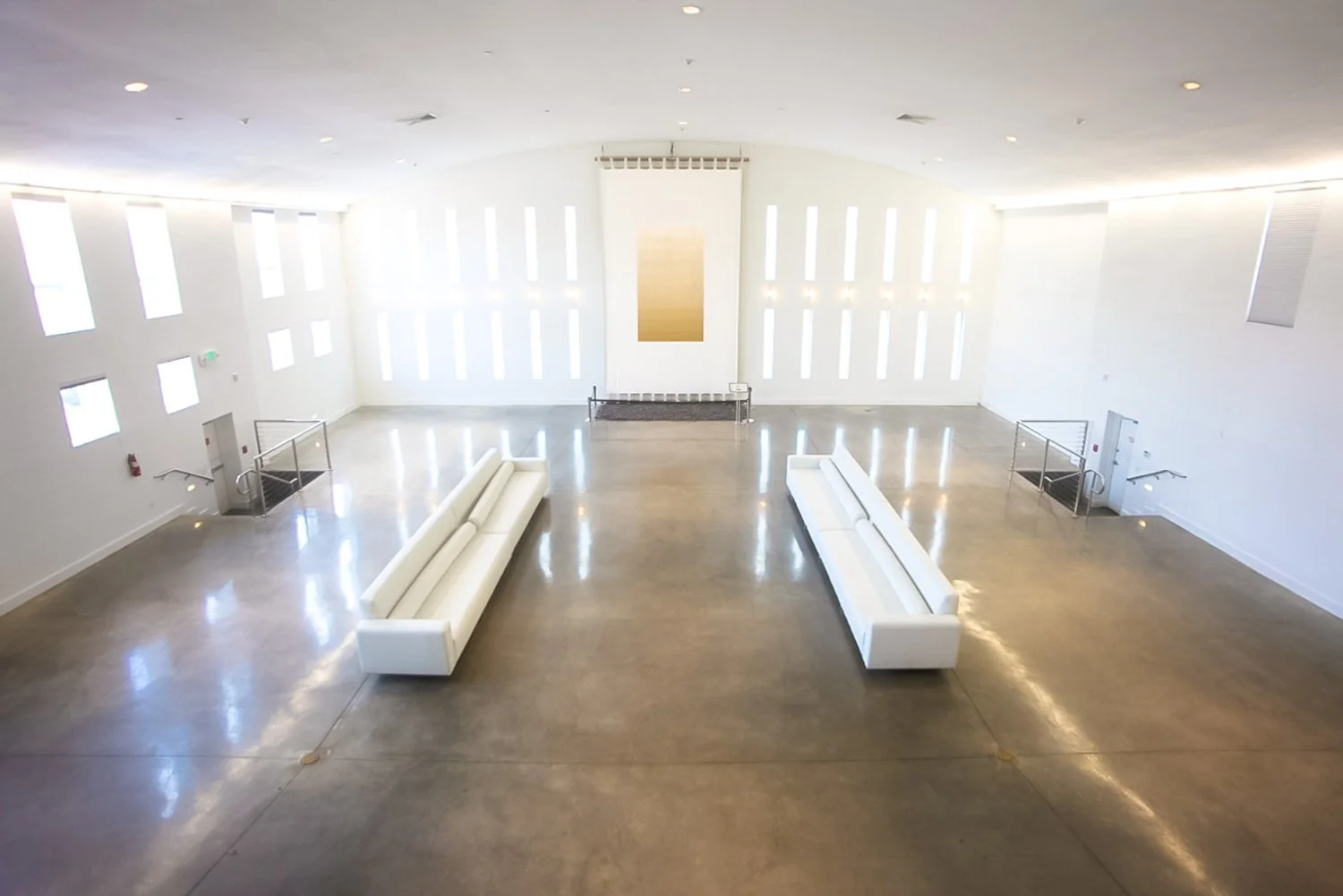 Empty, spacious modern hall with white walls, two long white sofas, a large artwork on the back wall, and multiple narrow windows allowing natural light.