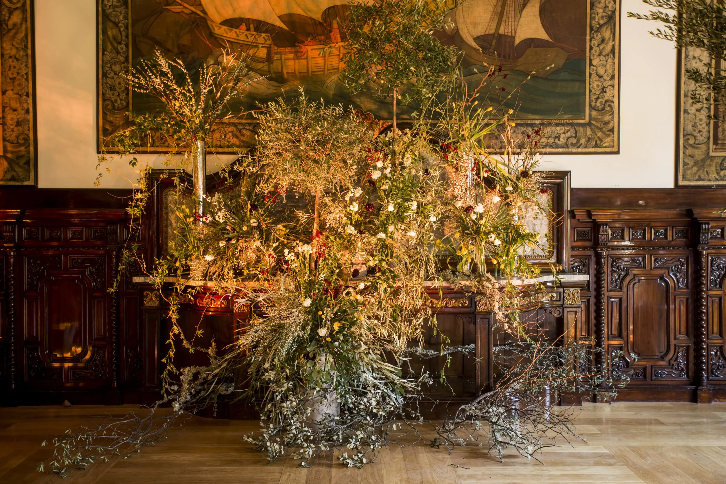 An elaborate flower and plant arrangement displayed in front of a large, ornate wooden cabinet, with a detailed framed painting or tapestry hanging on the wall behind, featuring a ship and the sea.