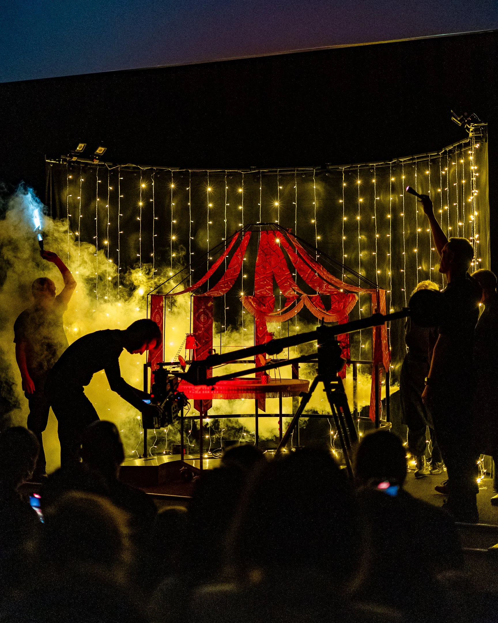 Nighttime scene of a circus tent with red and gold patterns, surrounded by string lights hanging from the top and fog or smoke at the base. Several silhouettes of people and a person holding a sparkler are visible in the foreground.
