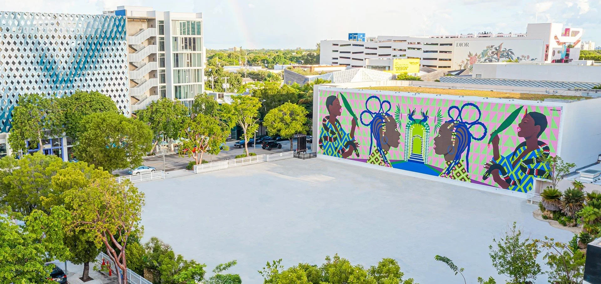Colorful street art mural depicting women with braided hair and traditional accessories on a building wall in an urban setting, with modern buildings, trees, and parked cars around.