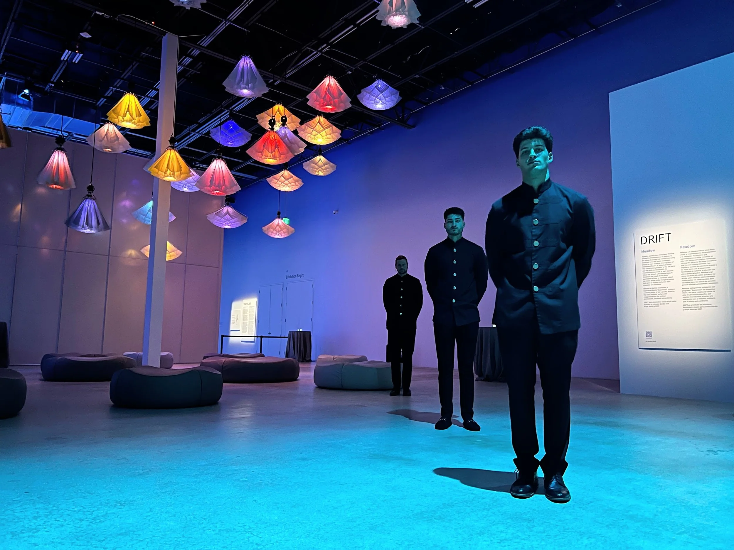 Three mannequins dressed in black stand in an art gallery with colorful umbrella-shaped light fixtures hanging from the ceiling. The gallery has a modern interior with a blue and purple lighting ambiance.