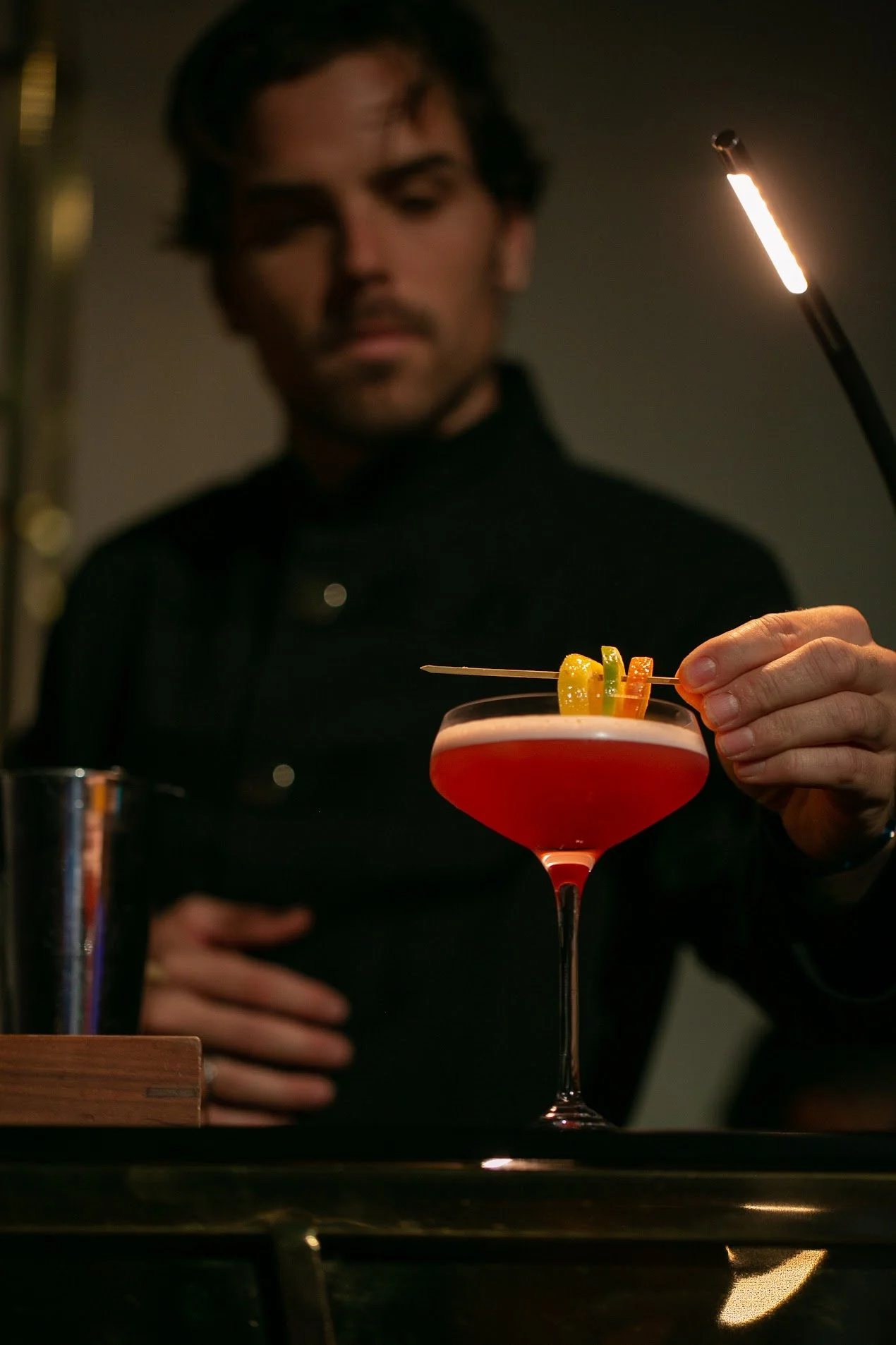 A bartender in black attire garnishing a red cocktail with an orange slice and a colorful garnish, under warm lighting.