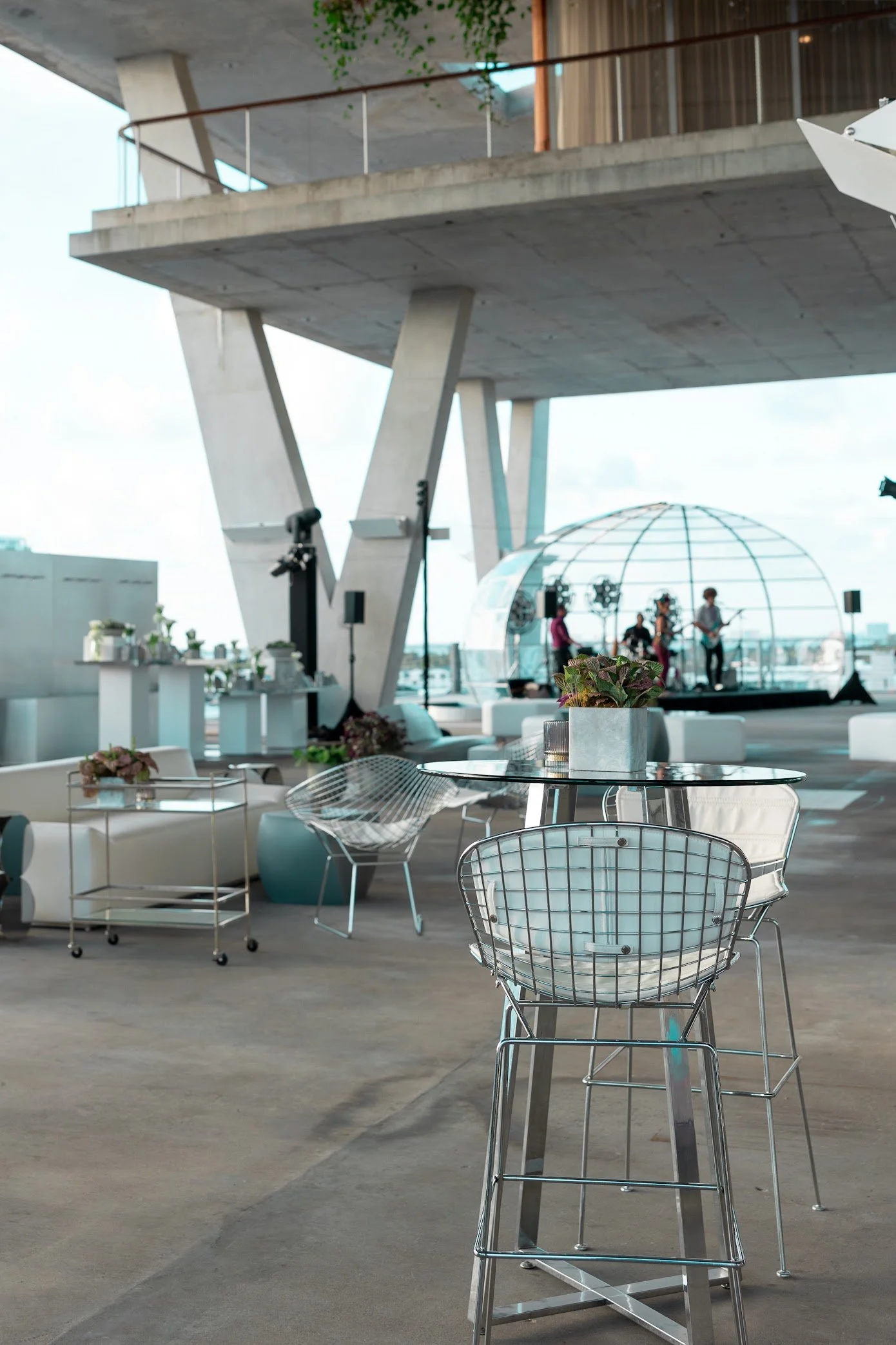Modern rooftop outdoor space with metal chairs, tables, and a stage with a band performing under a transparent geodesic dome. Urban buildings in the background.