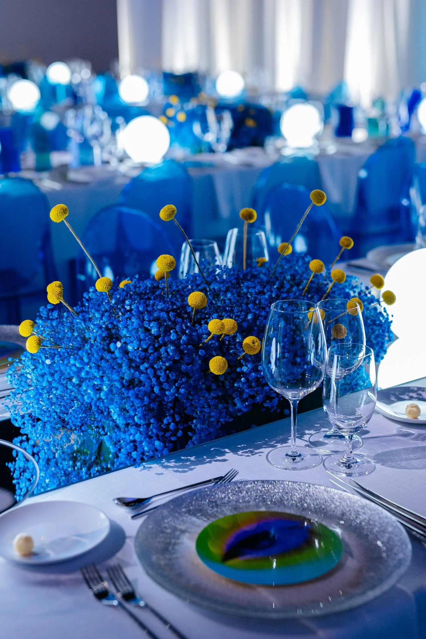Elegant banquet table decorated with a large blue floral centerpiece featuring small yellow flowers, surrounded by white tablecloths, wine glasses, plates, and silverware, with a blurred background of dinnerware and lighting.