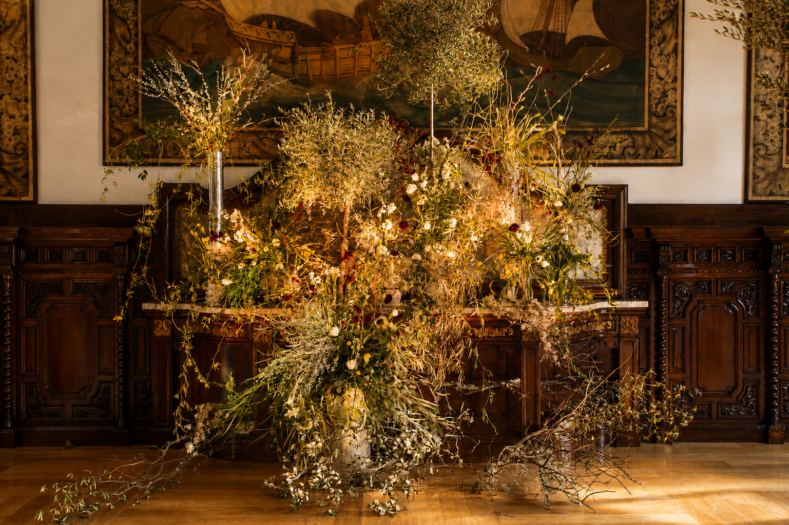 A large, elaborate floral arrangement with various green and white plants and flowers, set against a wooden-paneled wall with framed artwork.