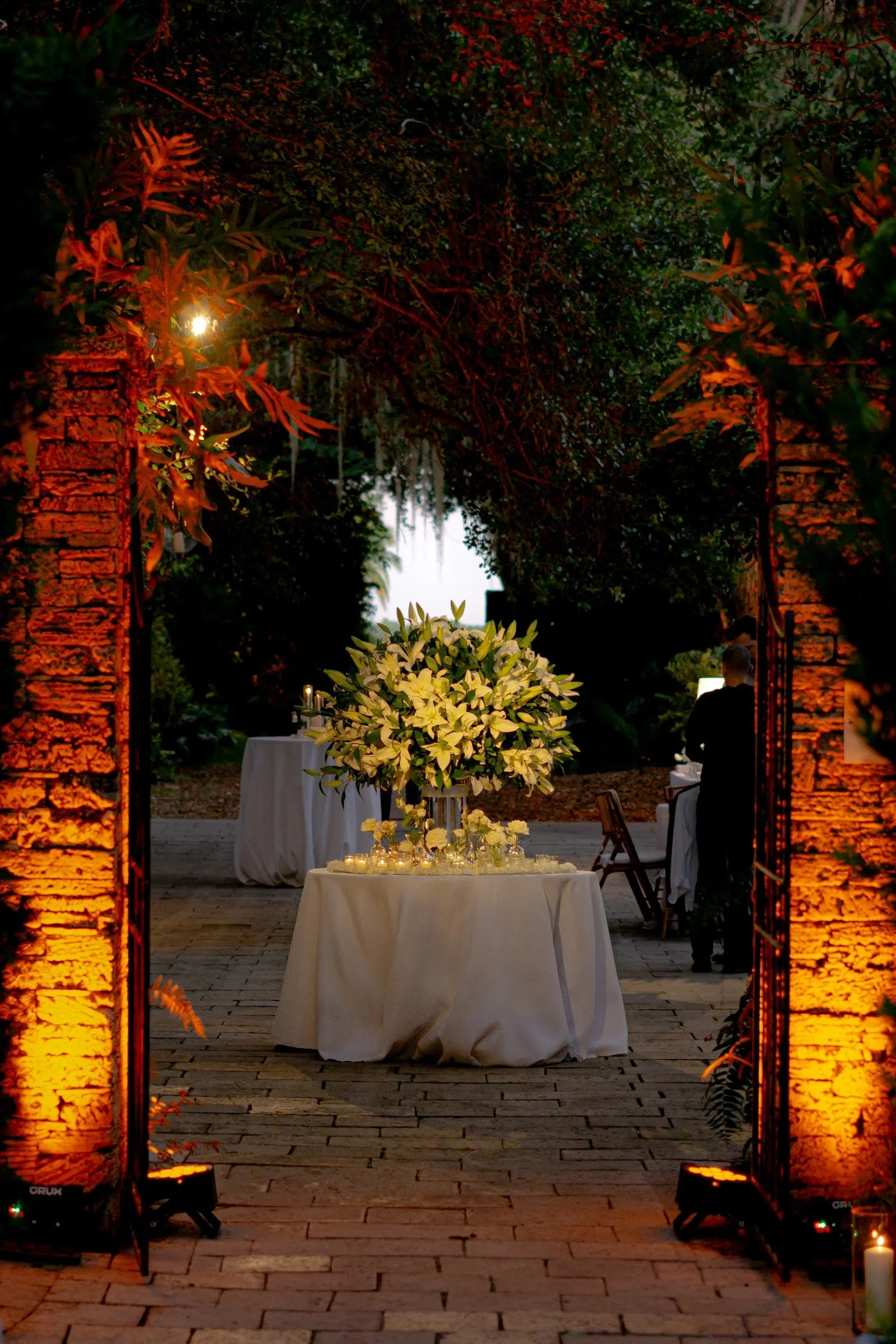 An outdoor evening event setup with a table decorated with white flowers and candles, framed by two illuminated brick pillars.