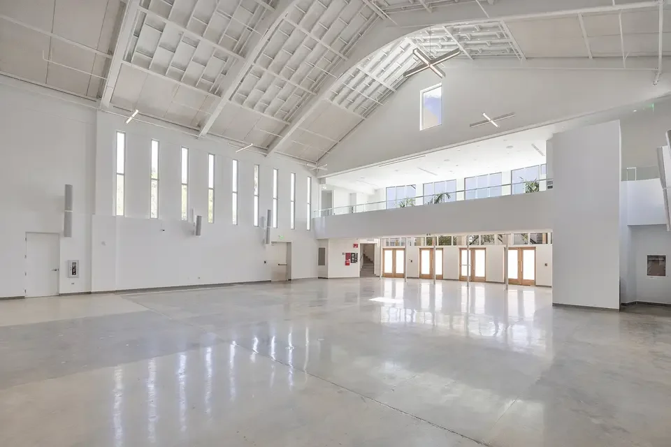 Interior of a modern, spacious building with white walls, high ceilings, large windows, and a glass railing on the second floor.