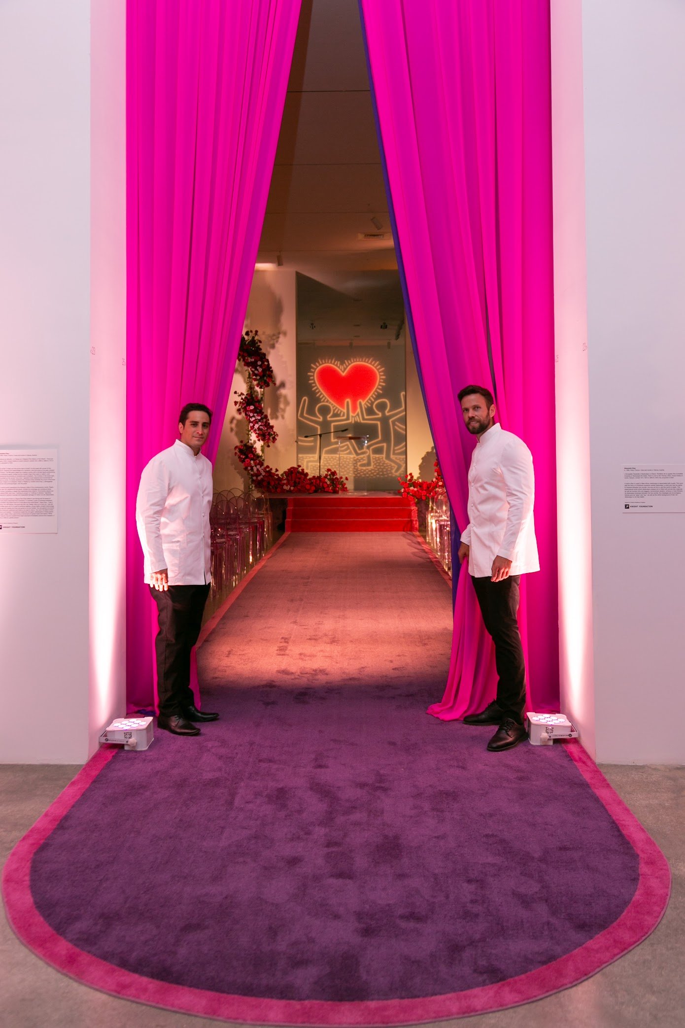 A pink and purple archway with pink curtains, leading into a room decorated for a romantic event with a heart-shaped neon sign and floral decorations, with two men in white shirts standing at the entrance.