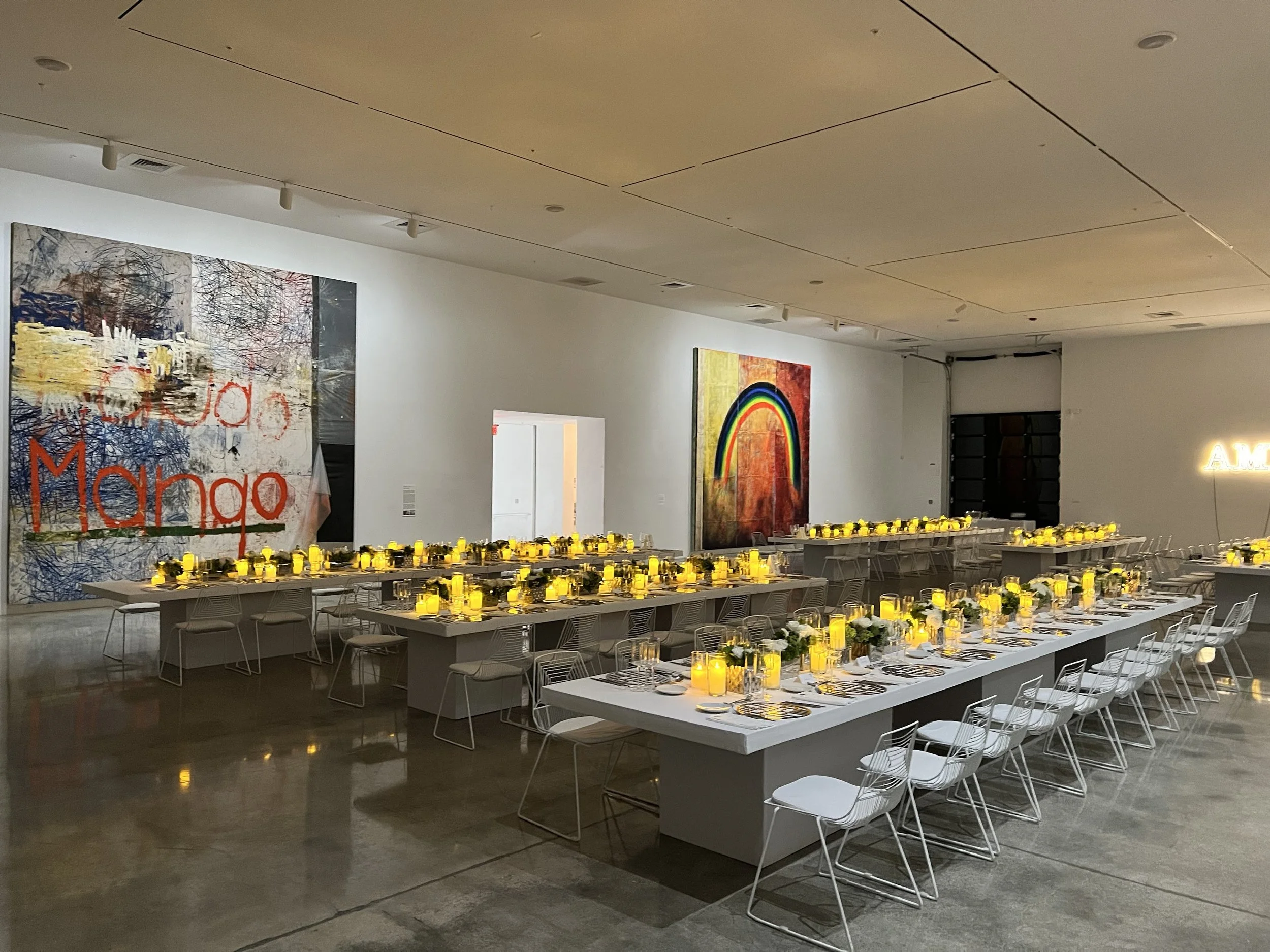 A large event space with long white tables set for a meal, decorated with yellow candles and flowers, and surrounded by white chairs. The walls feature colorful artwork, including a rainbow. The room is well-lit with ceiling lights.