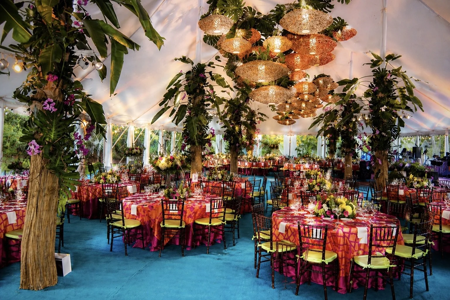 Decorated event tent with round tables covered in pinkish-orange tablecloths, black chairs with yellow cushions, floral centerpieces, and hanging lights and lanterns.