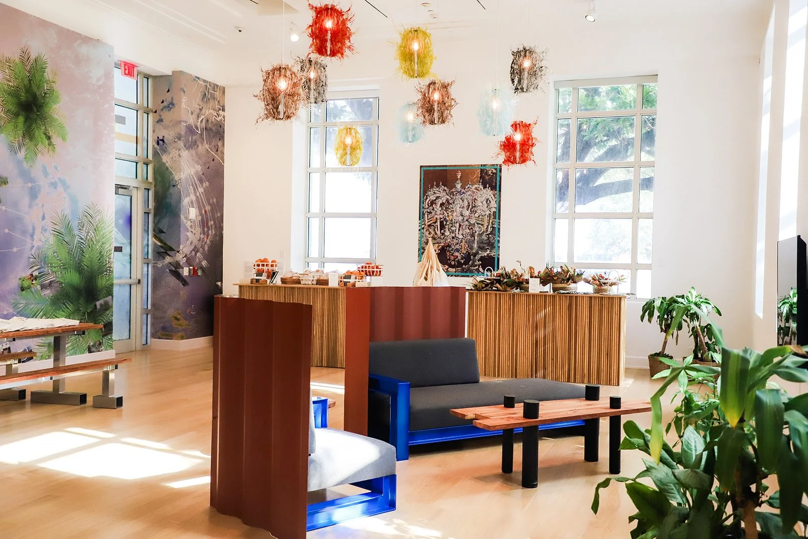 Interior of a modern, well-lit room with large windows, colorful hanging light fixtures, green plants, a dark gray couch, a wooden table, and decorative artwork on the wall.