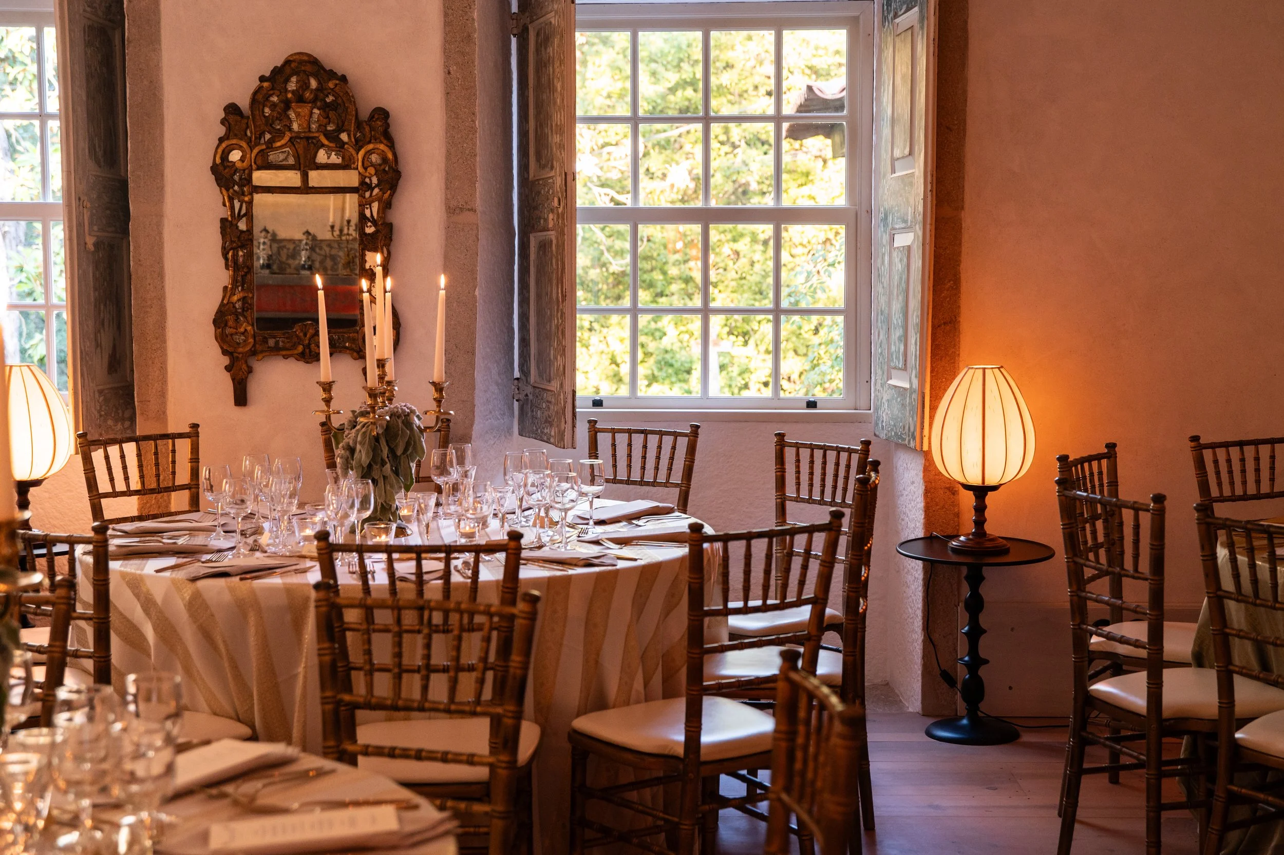 A dining room  at the Convento de Cabanas with a round table set with glassware and napkins, surrounded by wooden chairs. There is a window letting in natural light, and a table lamp with a lampshade on a small side table.