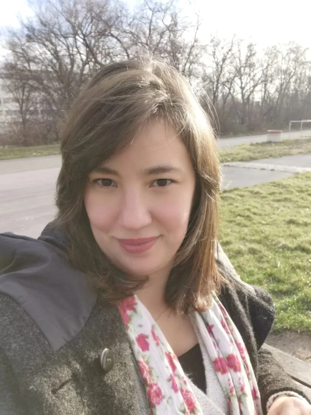 Nahaufnahme einer Frau mit braunen mittellangen Haaren, lächelnd, im Freien sitzend auf einer Parkbank bei Sonnenlicht, im Hintergrund Bäume und eine Wiese.