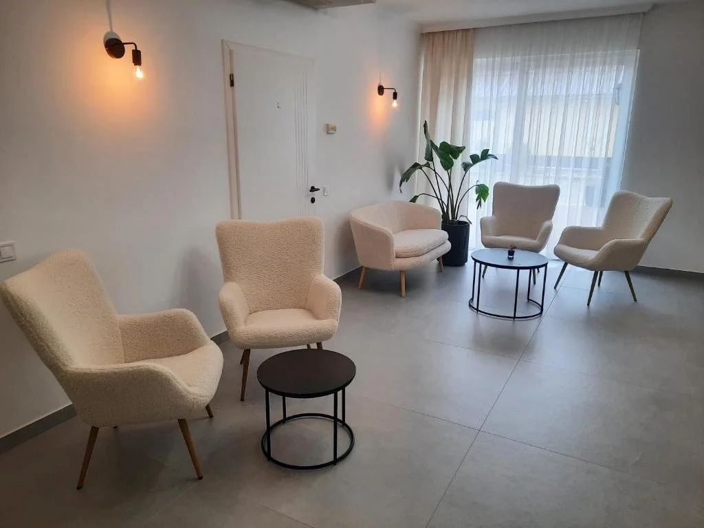 Gemütlicher Wartebereich mit sechs weißen Sesseln, zwei kleinen schwarzen Tischen, einem großen Pflanzkübel mit Pflanze, großer Fensterfront mit Vorhängen, weiße Wände und eingeschaltete Wandlampen.