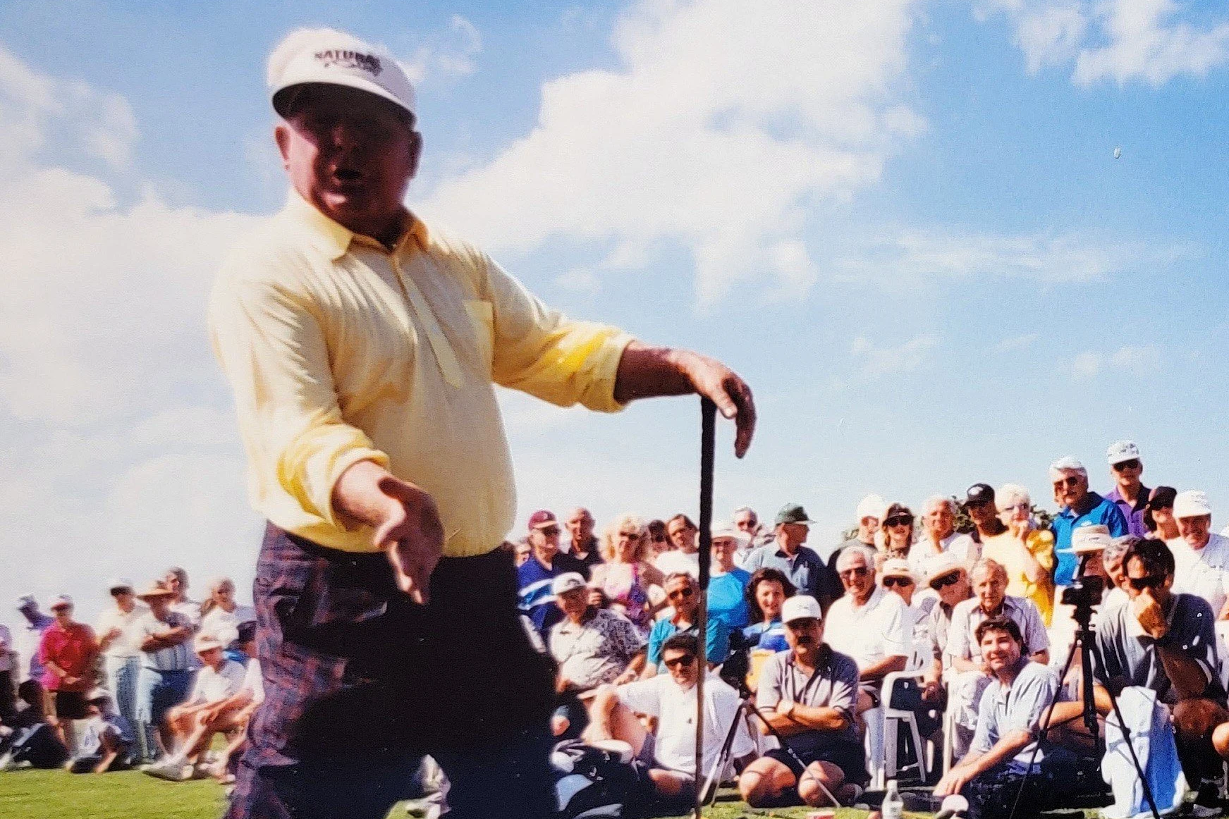 A man in a yellow shirt and plaid pants at a golf tournament, with a large crowd in the background watching him.