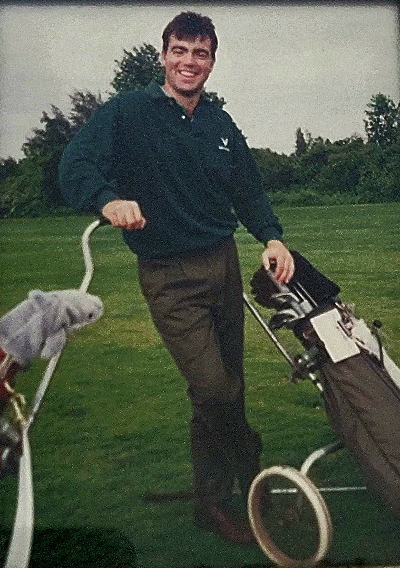 A young man smiling and holding golf clubs with a golf bag on a green golf course. There are trees in the background.