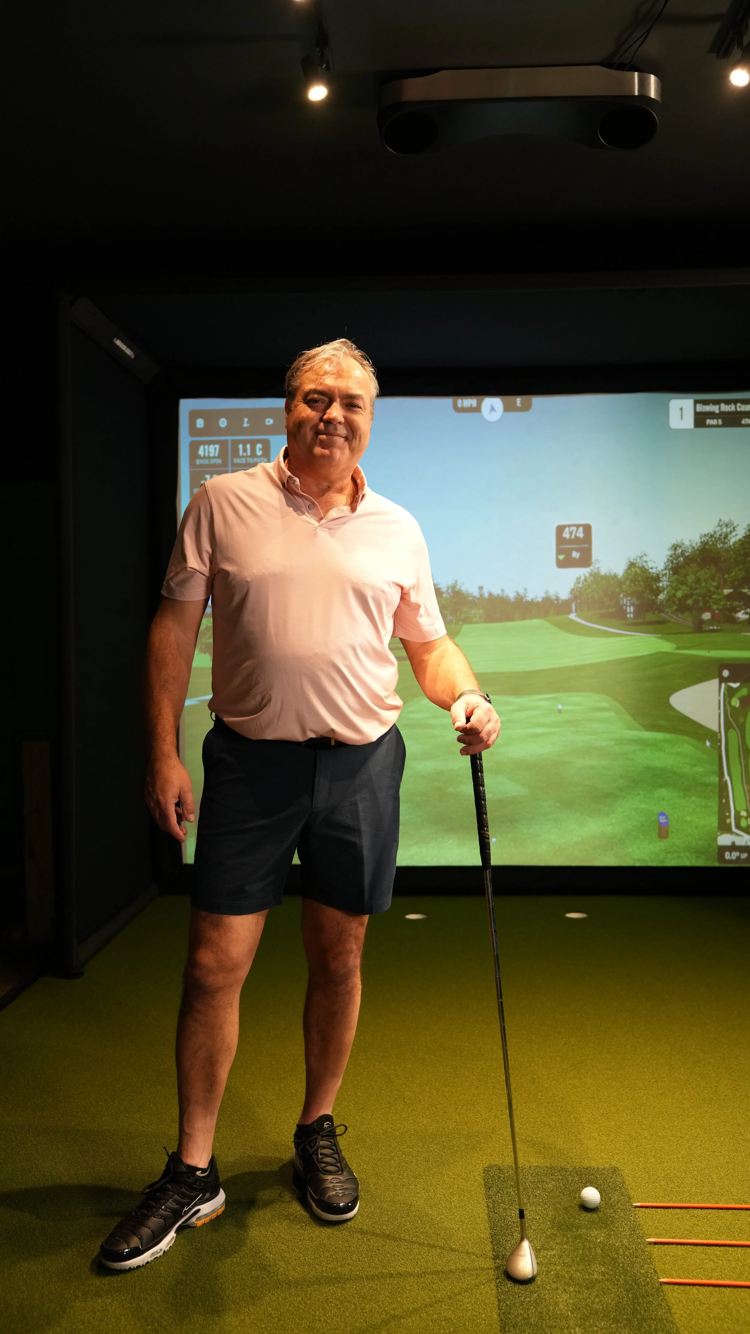 A man in a pink polo shirt and black shorts stands on an indoor golf simulator mat, holding a golf club with a golf ball positioned in front of him, in a room with a large screen displaying a virtual golf course.