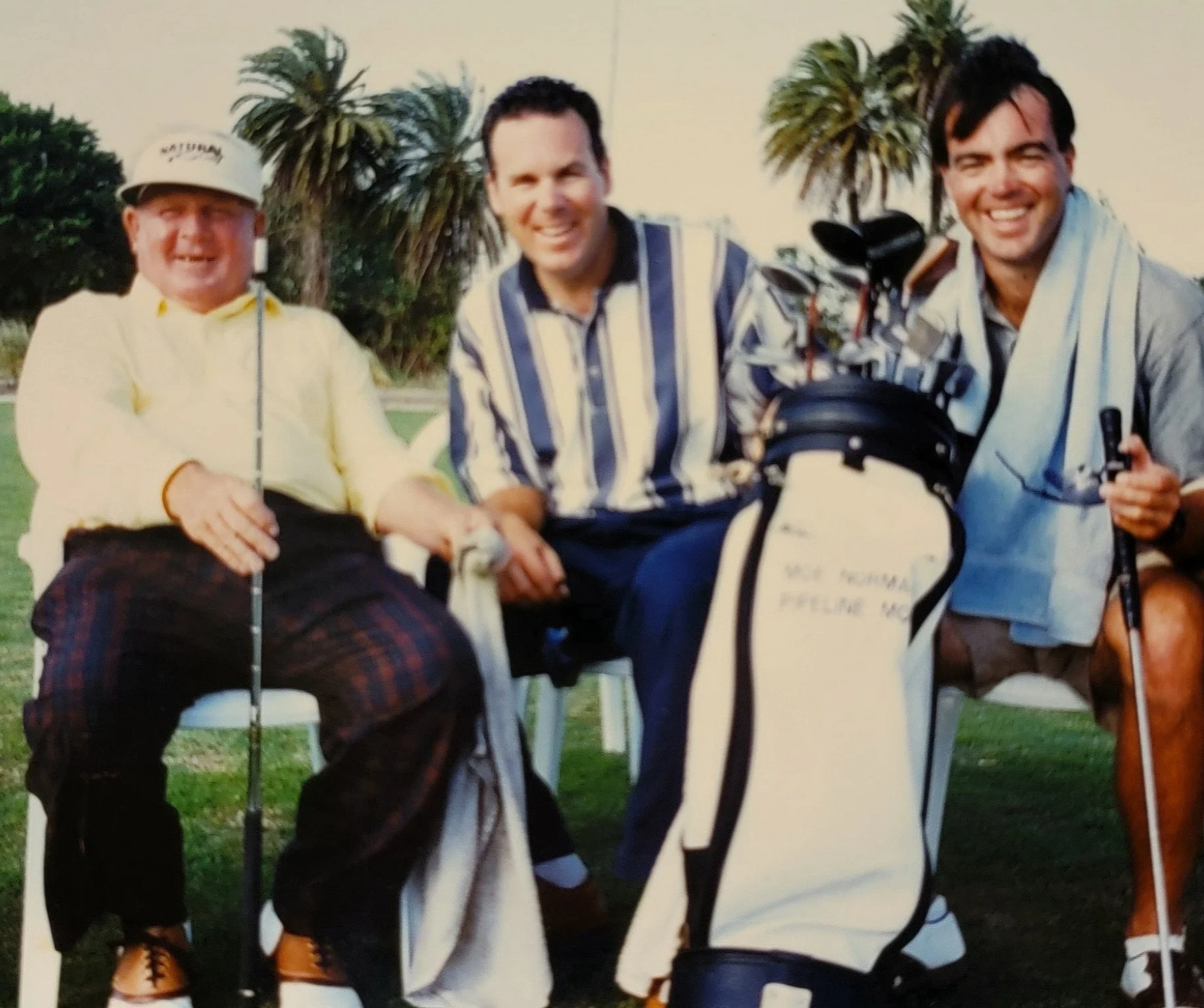 Three men sitting together outdoors, smiling at the camera, with palm trees in the background. The man on the left is wearing a light-colored jacket and a cap. The man in the middle is in a striped shirt, and the man on the right is wearing a light-colored shirt and is holding a golf club.
