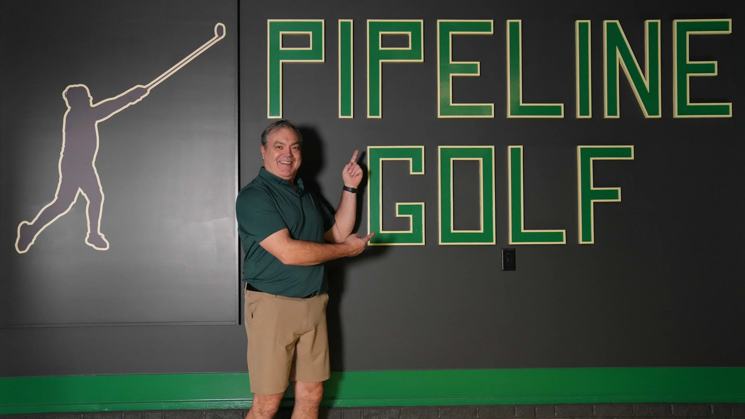 A man standing in front of a wall with the words 'PFEIFFER GOLF' in large green letters. The wall also features a silhouette of a golfer in mid-swing to the left of the text.