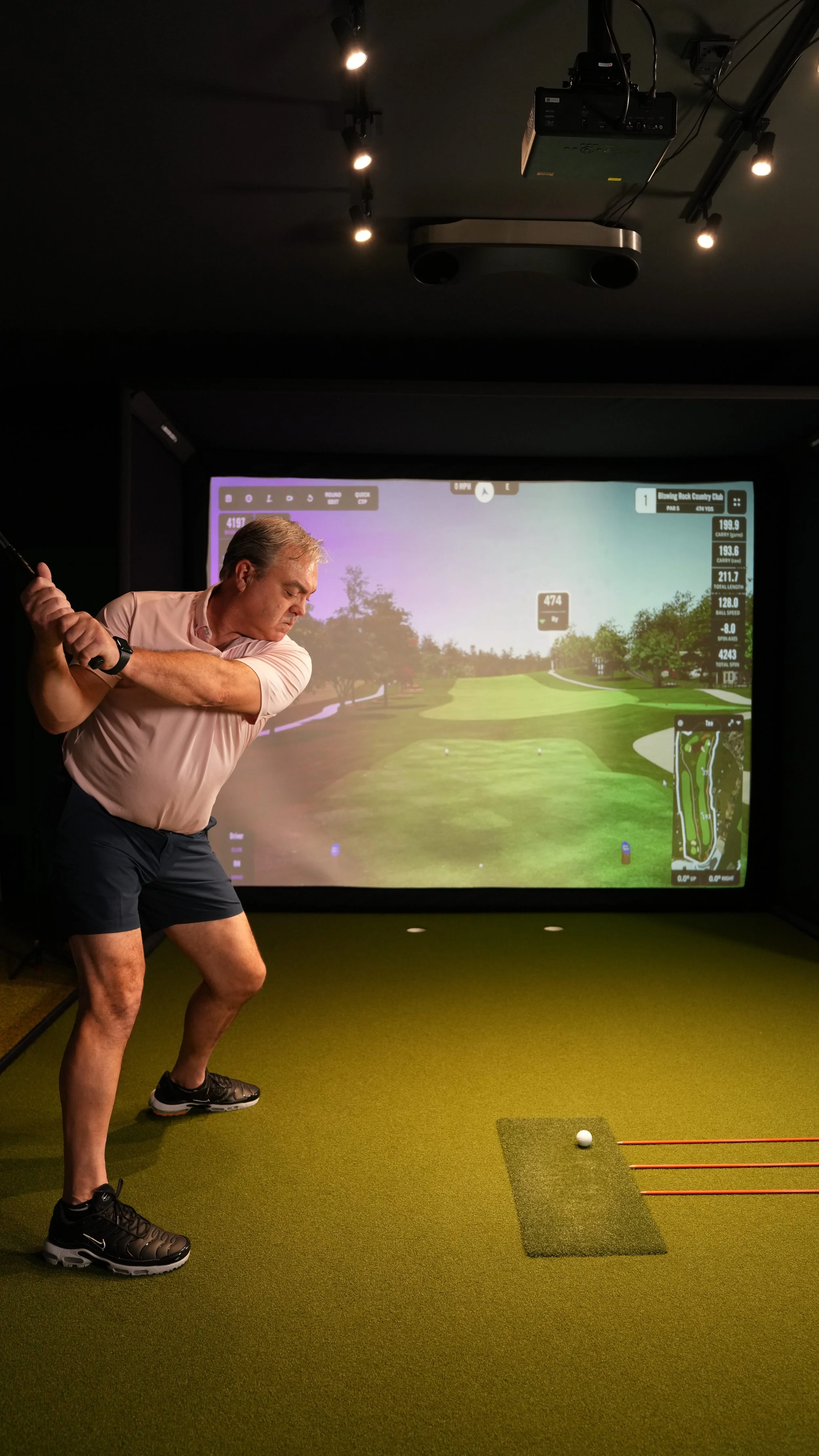 Man practicing golf swing in indoor golf simulator room with large screen displaying a golf course.