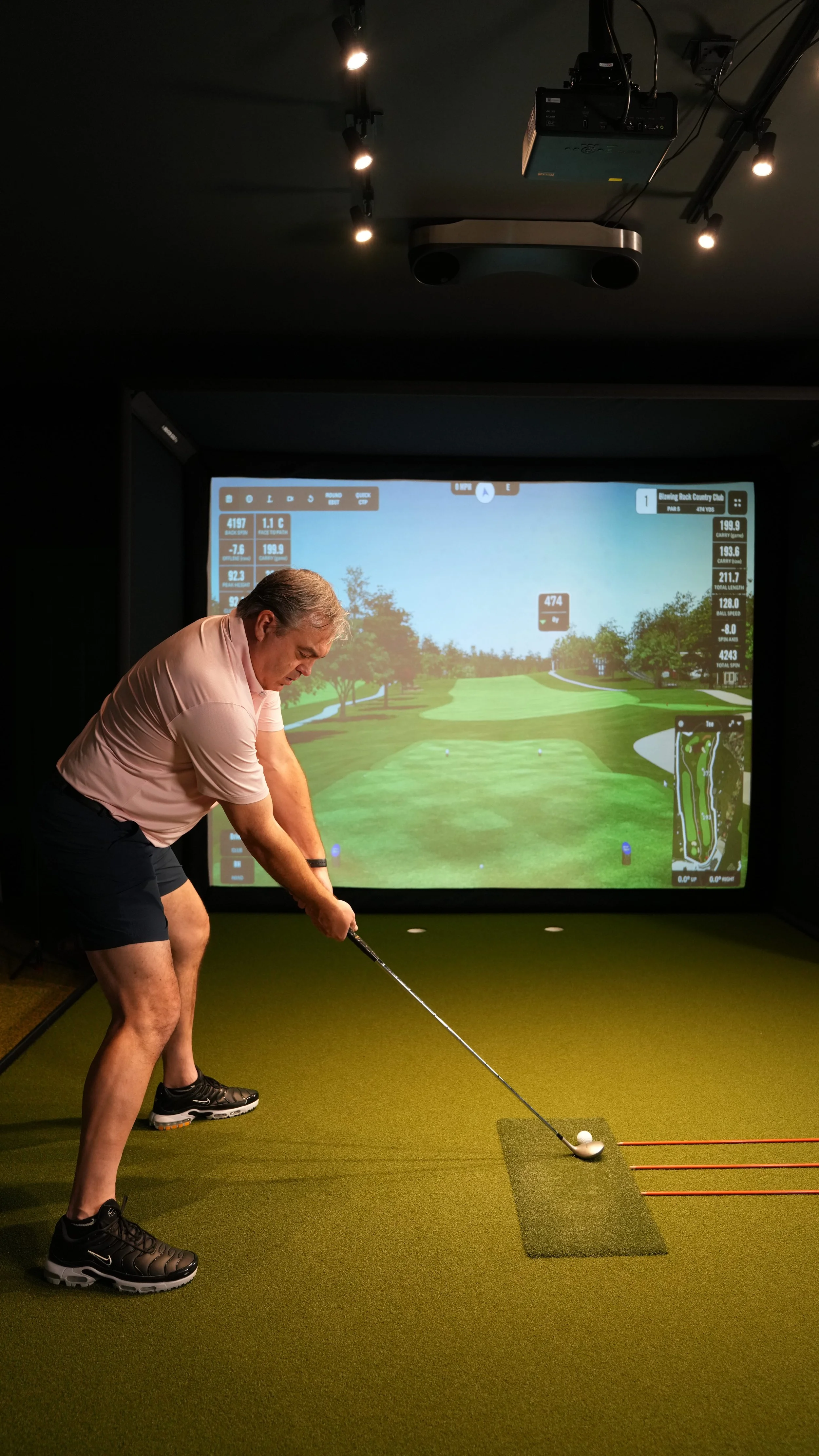 A man preparing to hit a golf ball on an indoor golf simulator with a large screen showing a virtual golf course in the background.