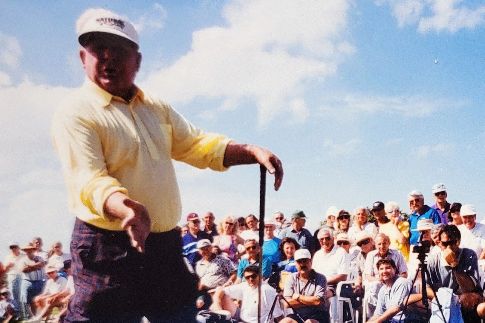 A man in a yellow shirt and cap standing in front of a large crowd of people outdoors on a sunny day.