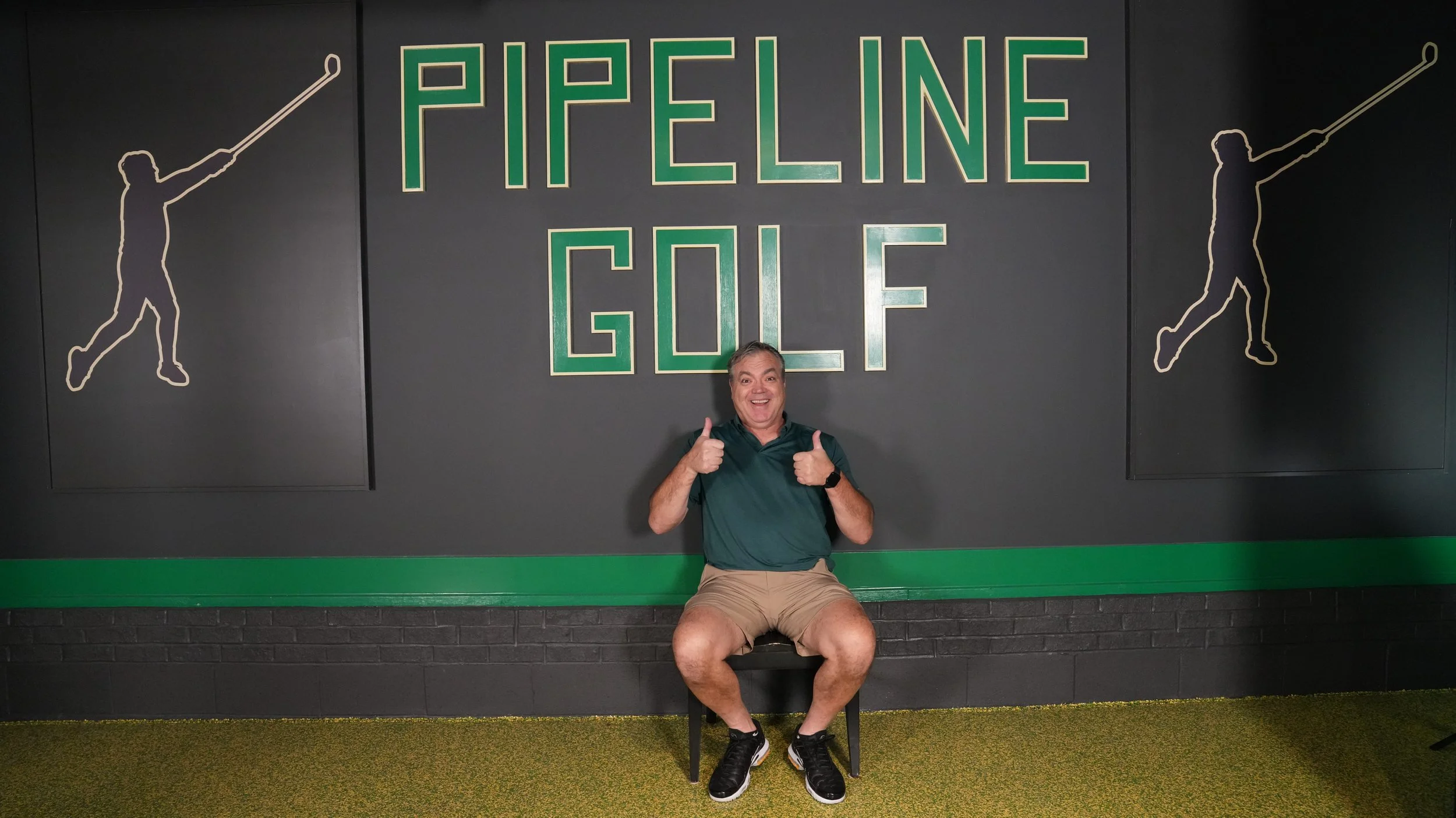 Man sitting on a bench in front of a large wall sign that reads 'PICKLE LIVE' with silhouettes of tennis players on either side of the text, giving thumbs up with both hands.