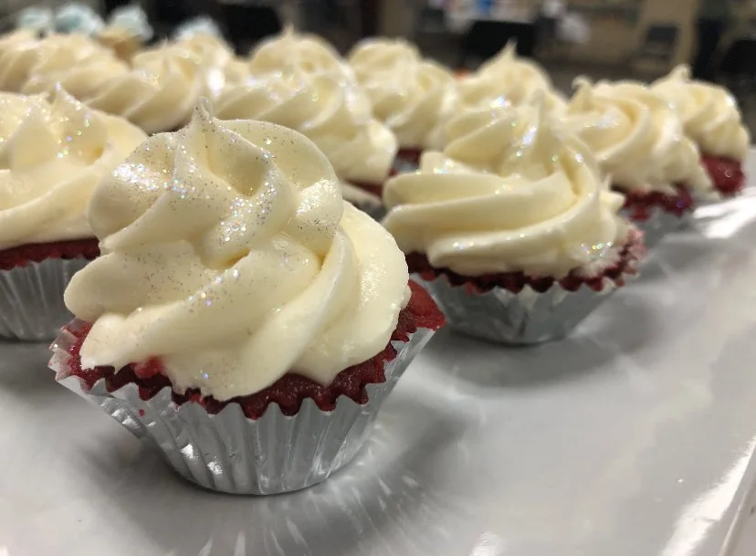 Close-up of red velvet cupcakes with cream cheese frosting and glitter sprinkles, lined up on a tray.