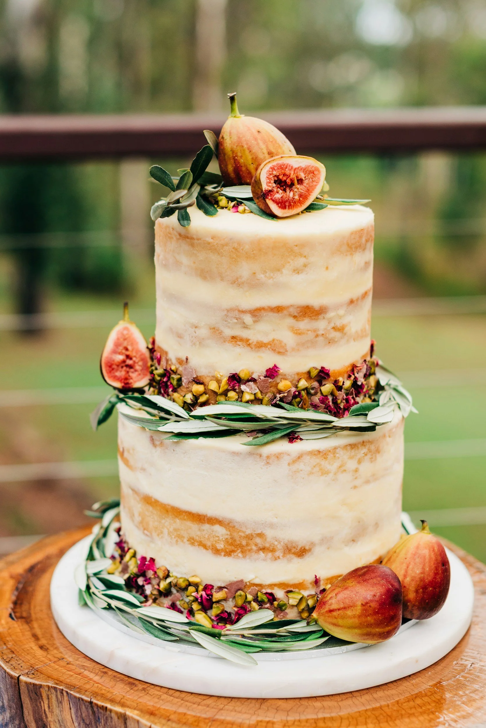 A two-tier semi-naked wedding cake decorated with figs, almonds, and greenery, placed on a round wooden slab.