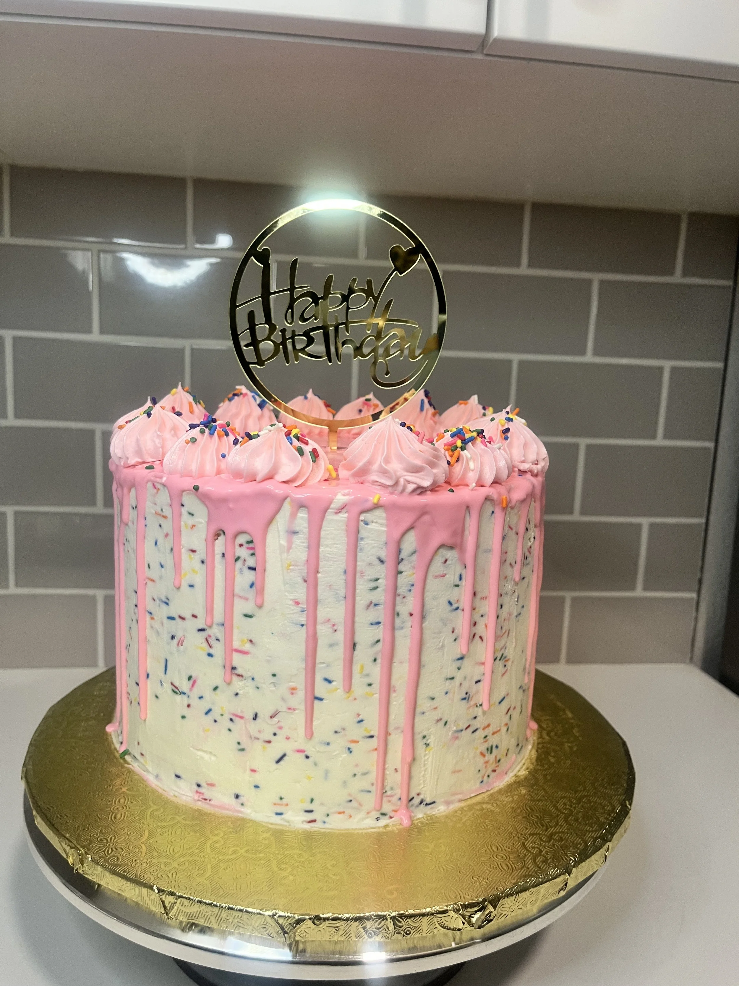 Pink and white birthday cake with rainbow sprinkles, pink icing drips, pink whipped cream dollops on top, and a "Happy Birthday" topper.