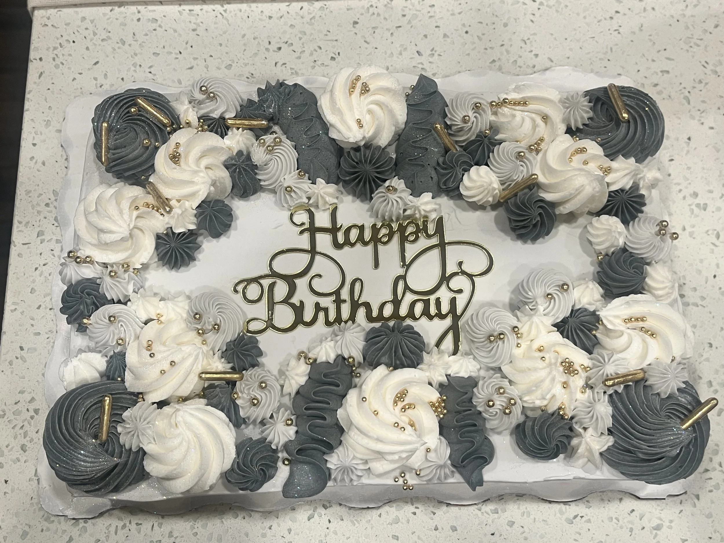 A rectangular birthday cake decorated with white, black, and gray whipped cream, gold beads, and a gold 