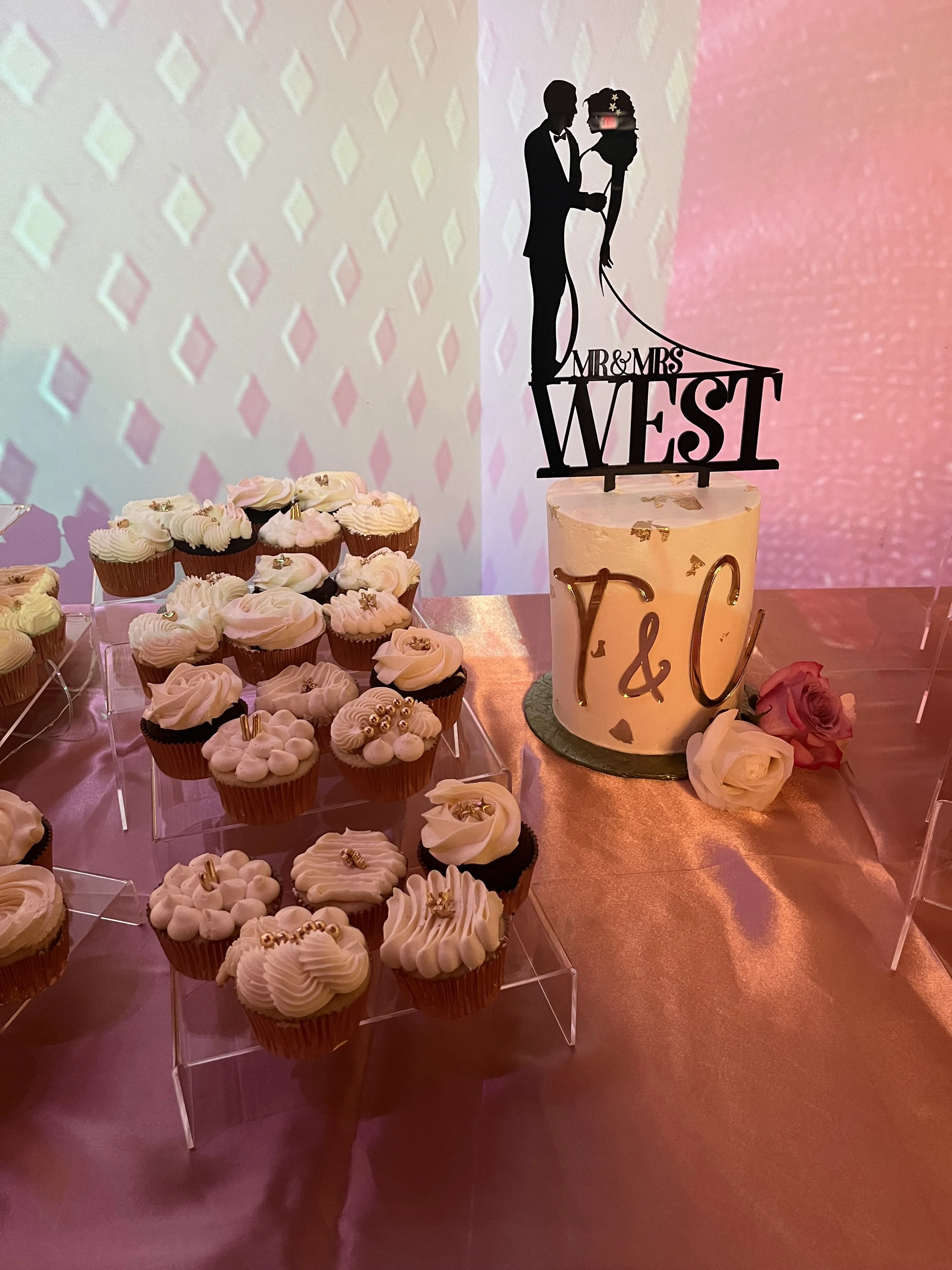A wedding cake with a silhouette topper of a couple holding hands, and cupcakes decorated with cream and gold accents. The cake has 'Y & C' written on it, and there are pink and white roses beside it. The background features a projected pattern and p
