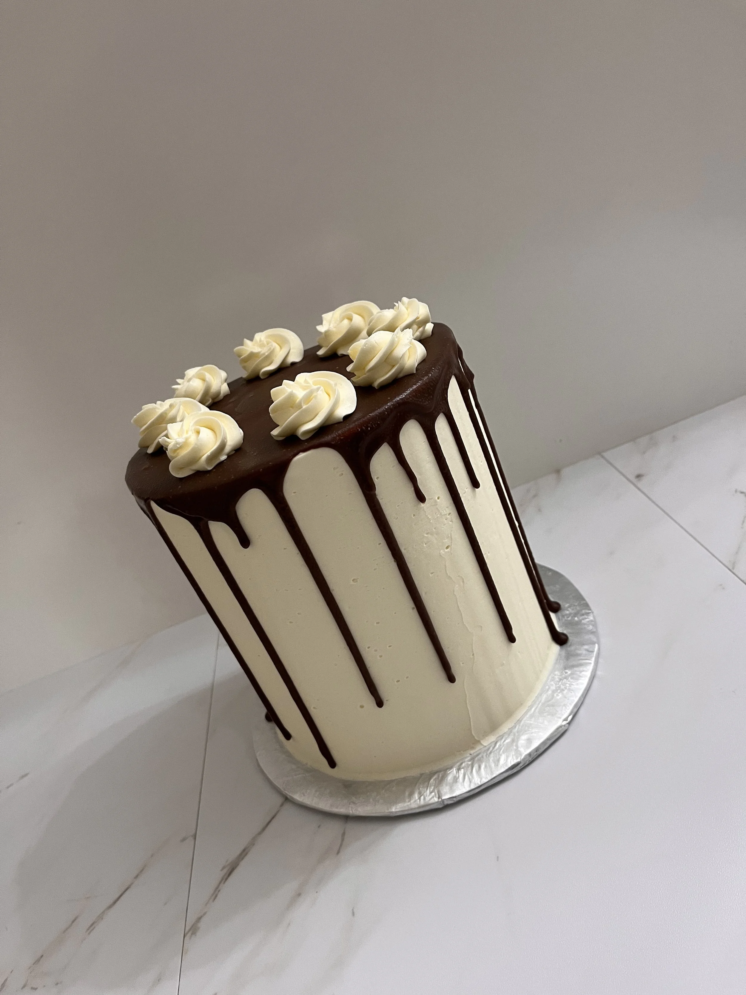 A tall layered cake with white frosting, decorated with white frosting rosettes on top, and chocolate ganache dripping down the sides, placed on a silver cake board.