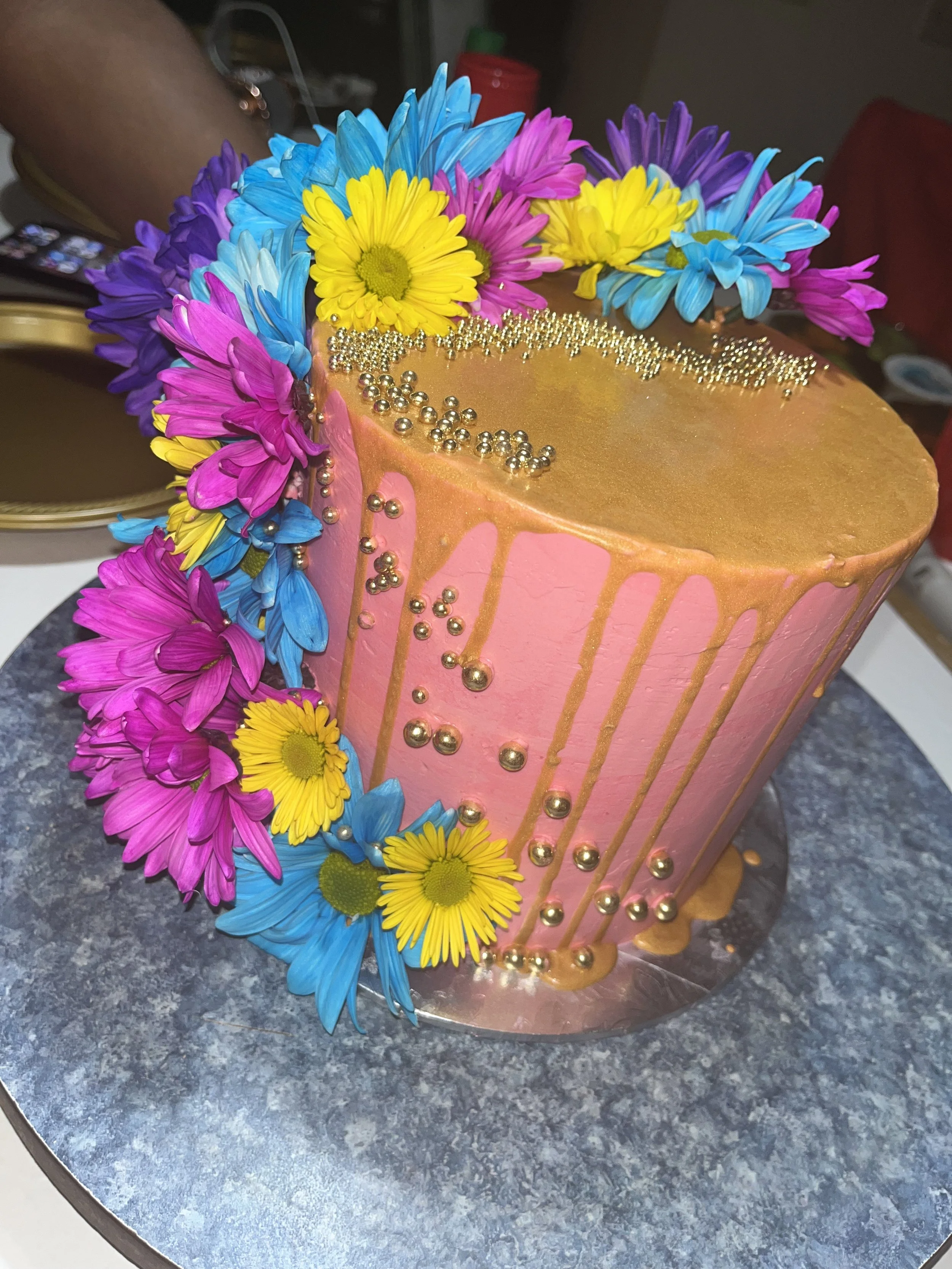 A pink cake decorated with colorful flowers, gold drip icing, and small gold and silver edible beads.