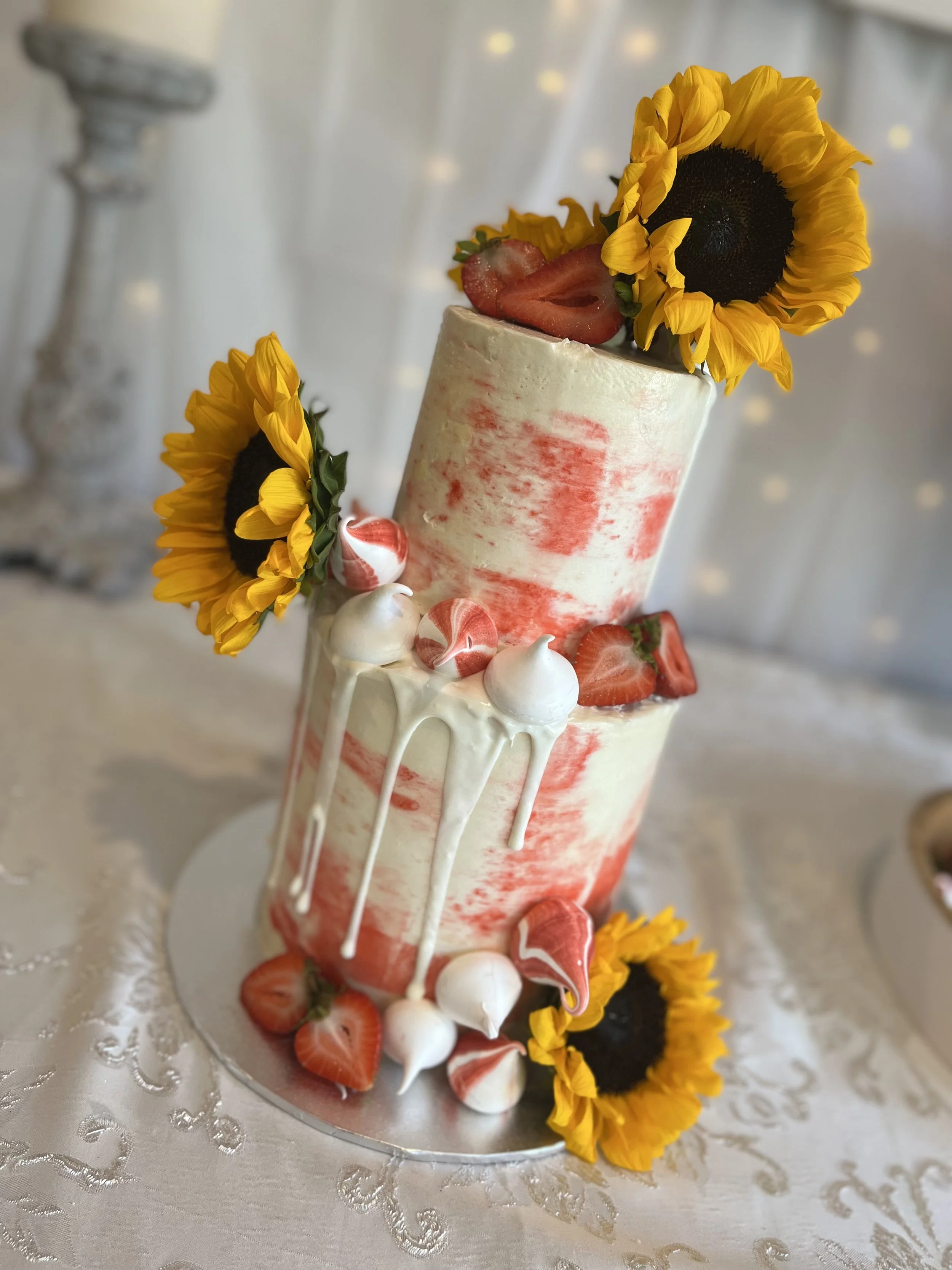 A two-tiered wedding cake decorated with strawberries, white and pink meringues, white chocolate drips, artificial sunflower flowers, and a strawberry on top, set on a decorative tablecloth.