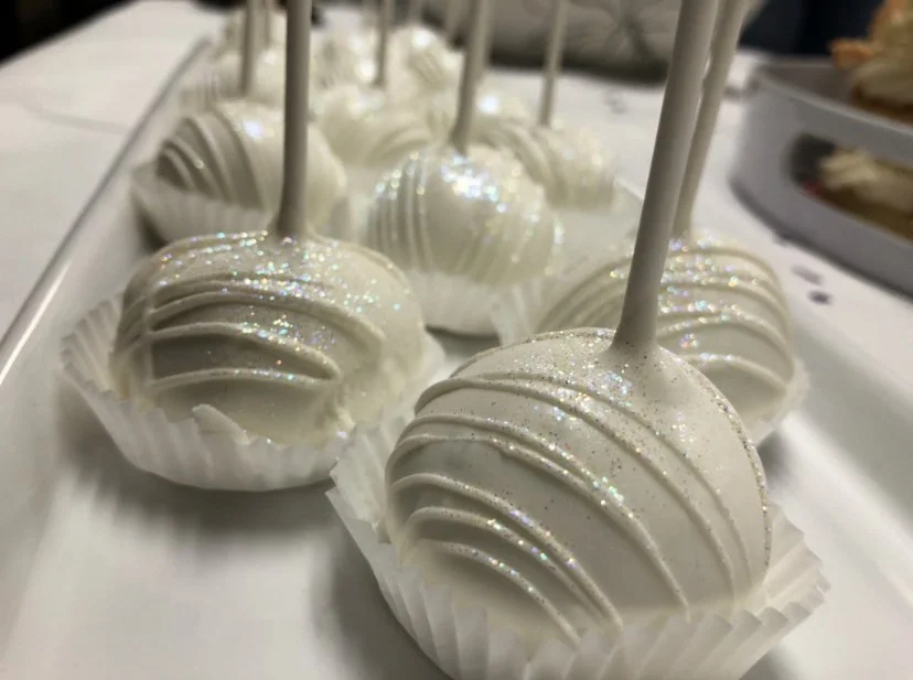 Close-up of white cake pops decorated with shimmer and placed in white paper cups on a dessert tray.