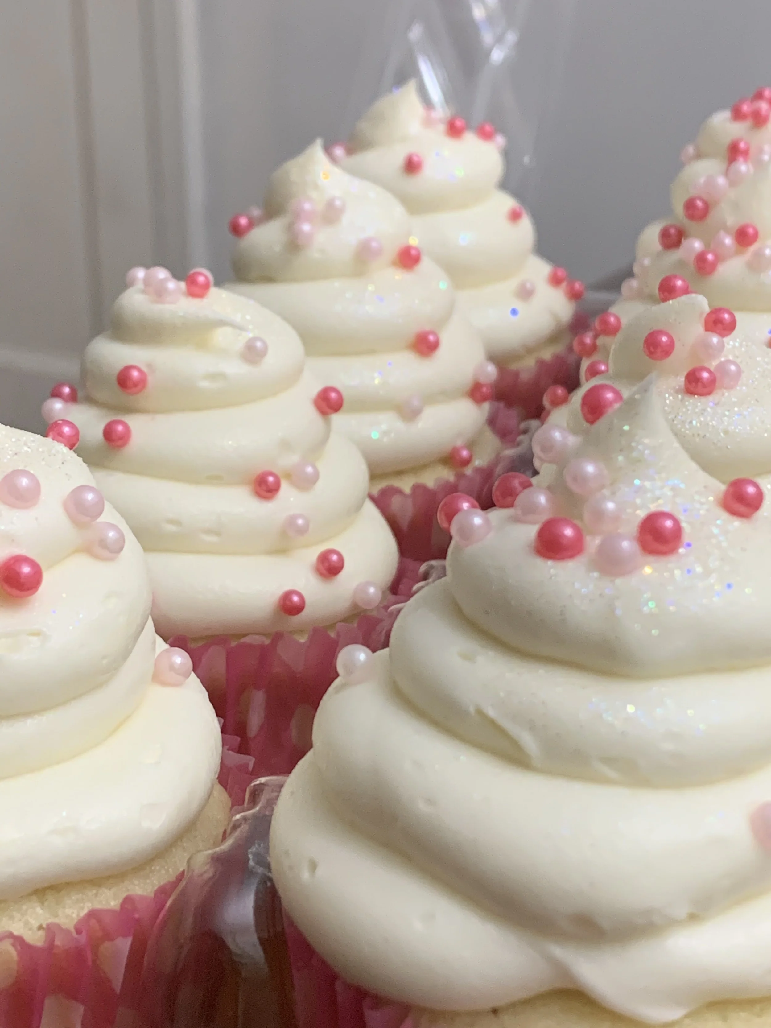 Close-up of decorated cupcakes with white frosting, pink and red pearl sprinkles, and glitter, in pink cupcake liners.