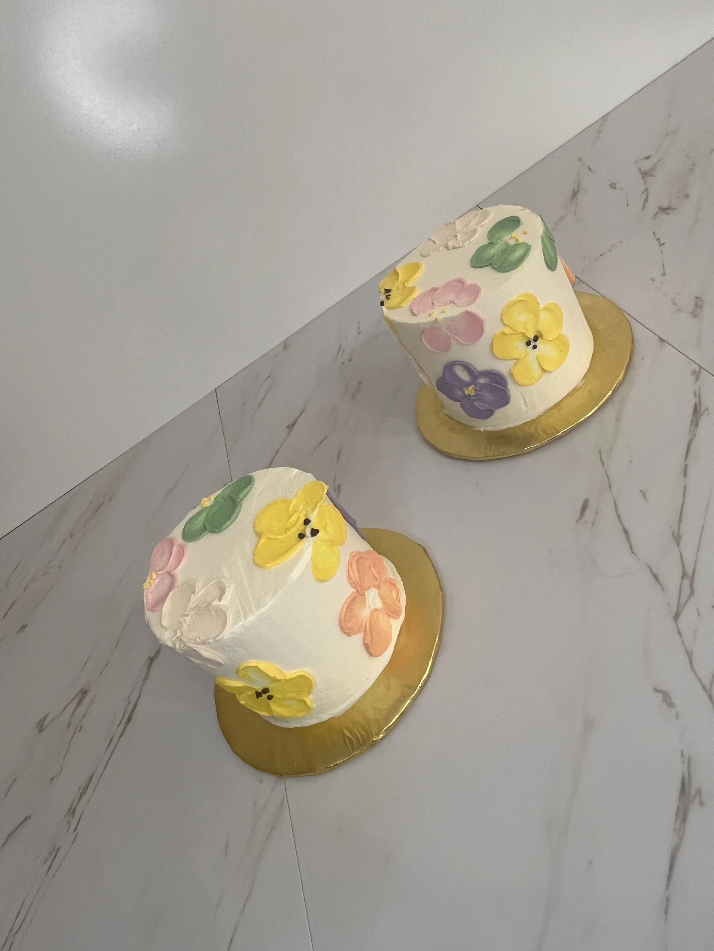 Two white cakes decorated with pastel-colored flowers and yellow, pink, purple, and green accents on a gold cake board, placed on a marble surface.