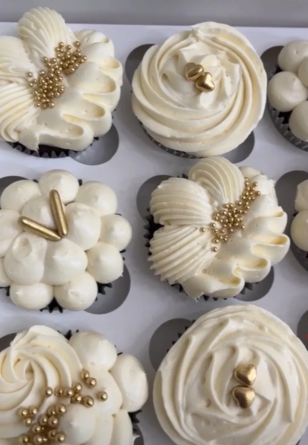 Assorted cupcakes with white frosting, decorated with gold-colored pearls, hearts, and metallic figurines.