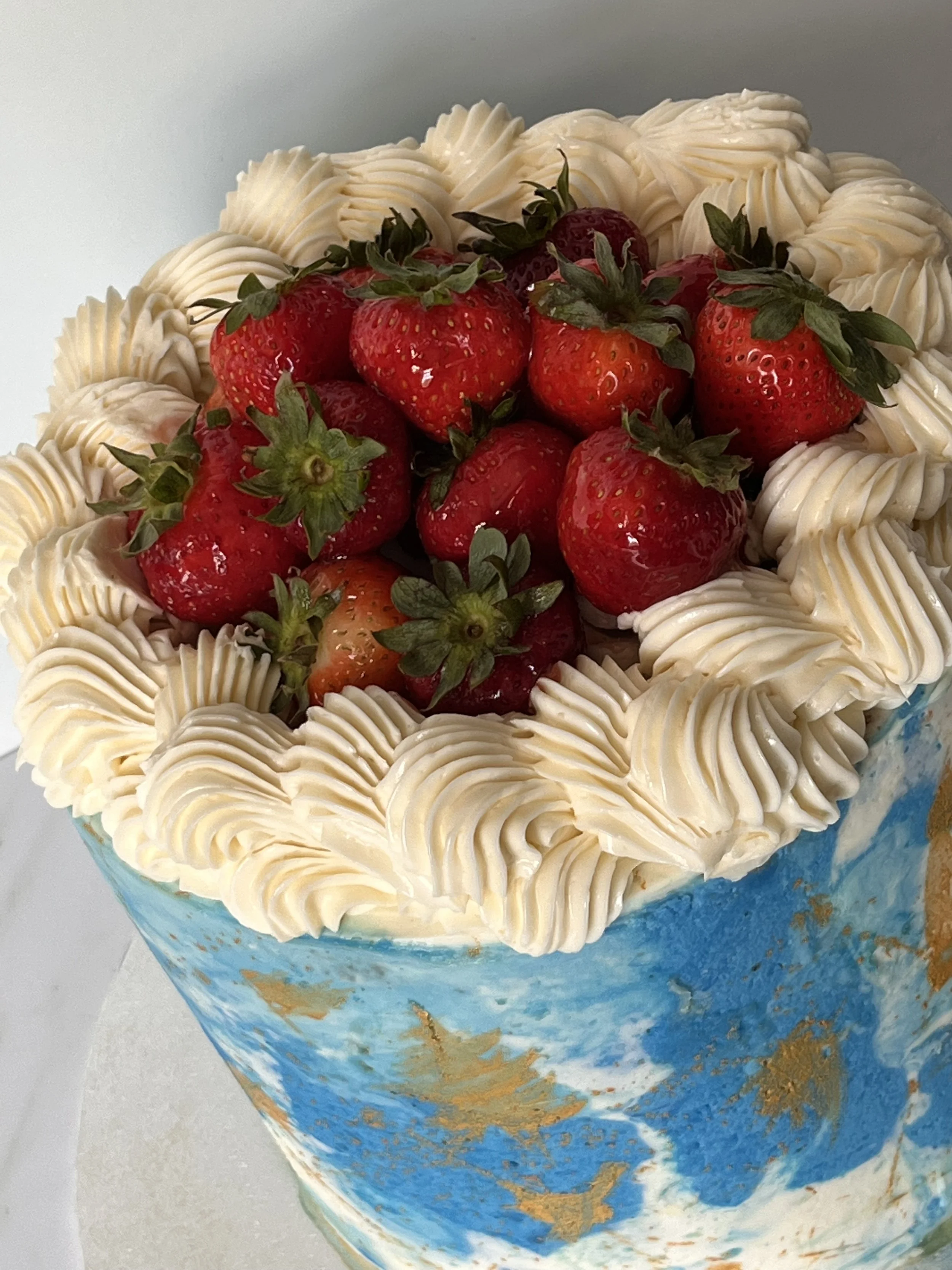 A cake decorated with strawberries on top and white frosting piped around the edges, with a blue and gold marbled design on the sides.