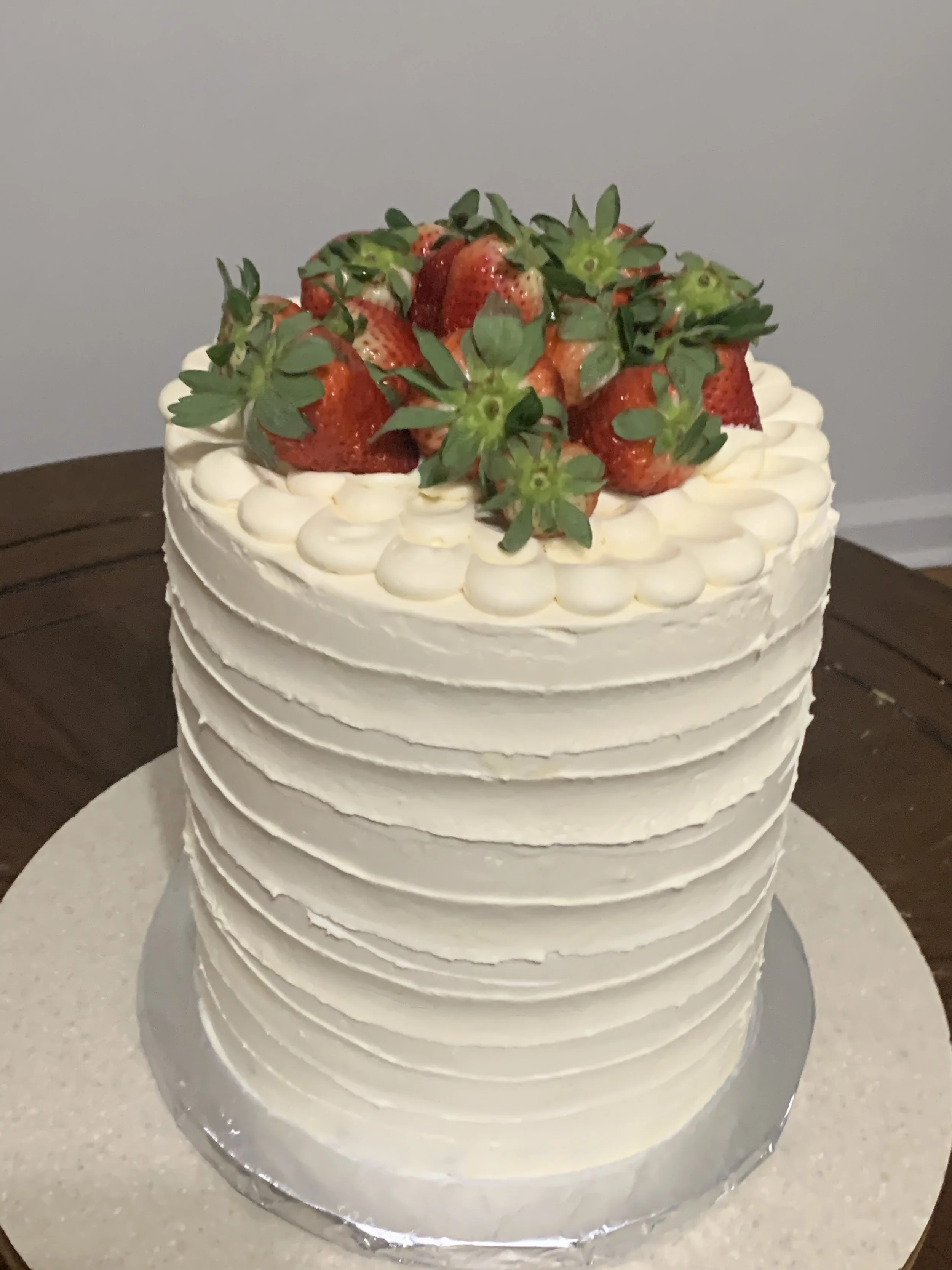 A tall, multi-layered white cake decorated with fresh strawberries and green leaves on top, with piped white frosting around the edges.