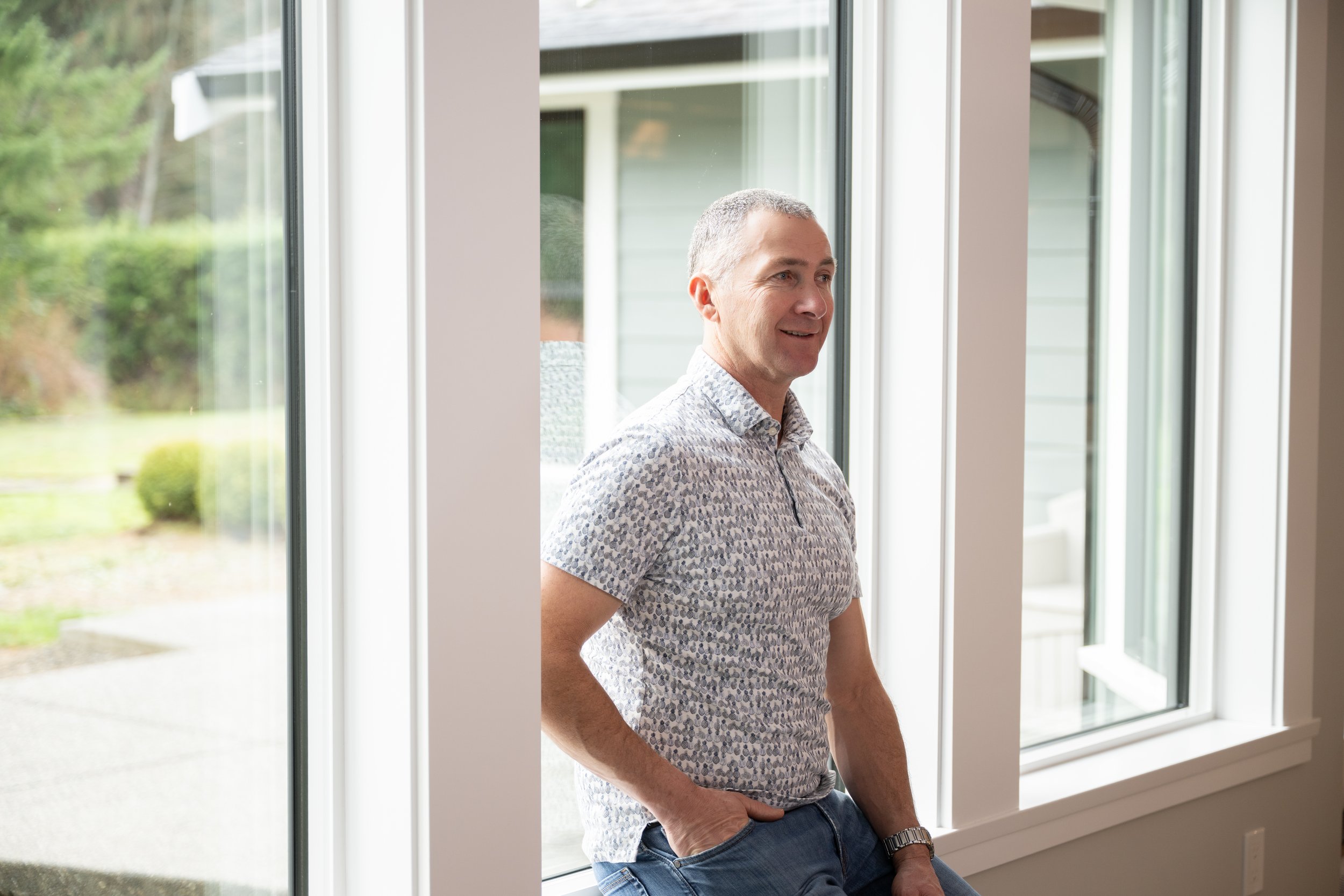 A middle-aged man with short gray hair standing indoors near a large window, smiling as he looks outside. He is wearing a short-sleeved patterned shirt and jeans.