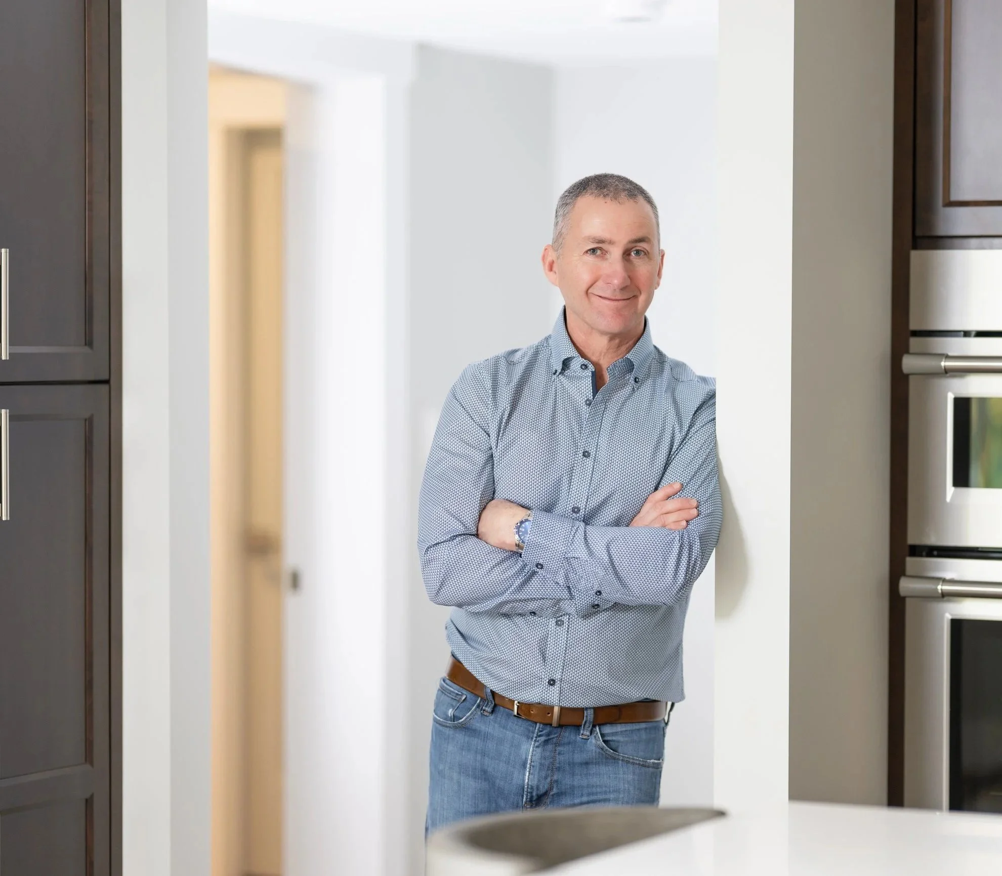 A middle-aged man with short gray hair, wearing a light blue patterned shirt and jeans, is standing in a modern kitchen with his arms crossed, smiling slightly, next to a wall.