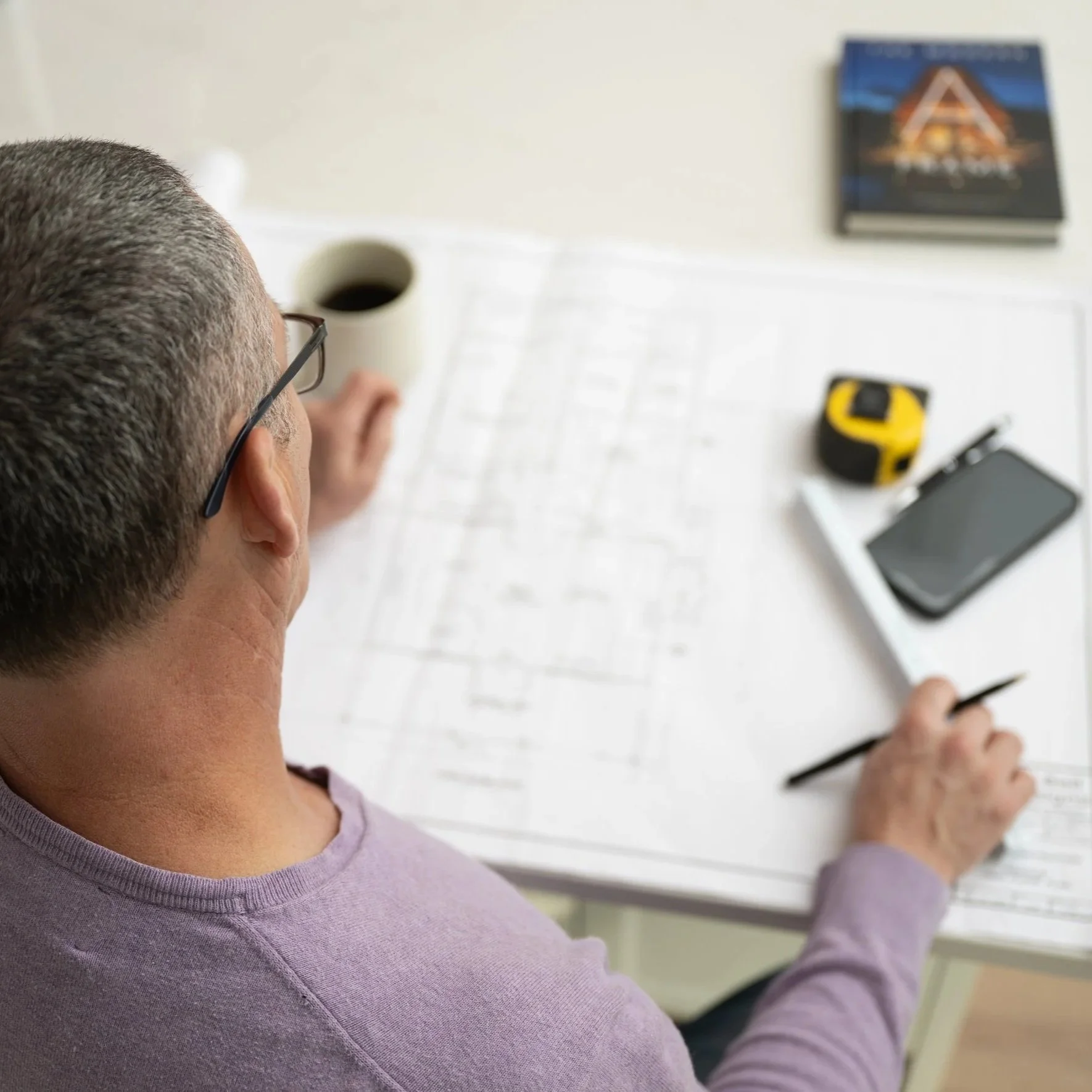 A person with short hair and glasses, sitting at a desk, looking at architectural plans. The desk has a cup of coffee, a measuring tape, a smartphone, and a blueprint.