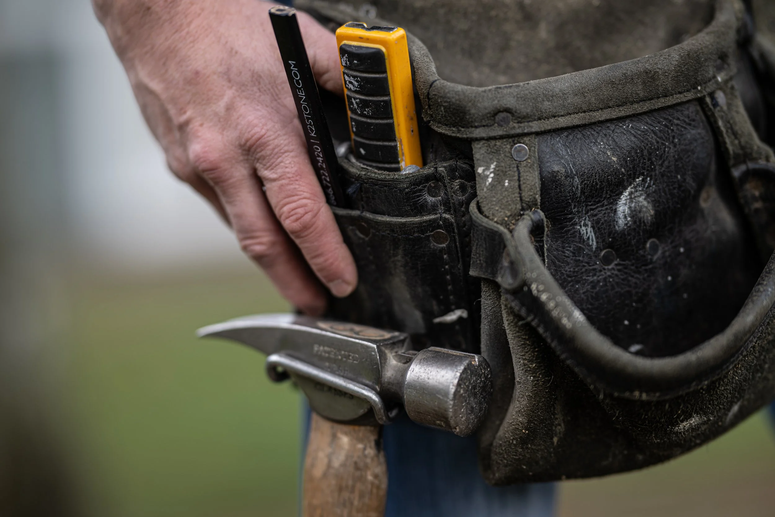 A person placing a chisel into a leather tool pouch attached to their waist, with a hammer hanging below the pouch, outdoors.