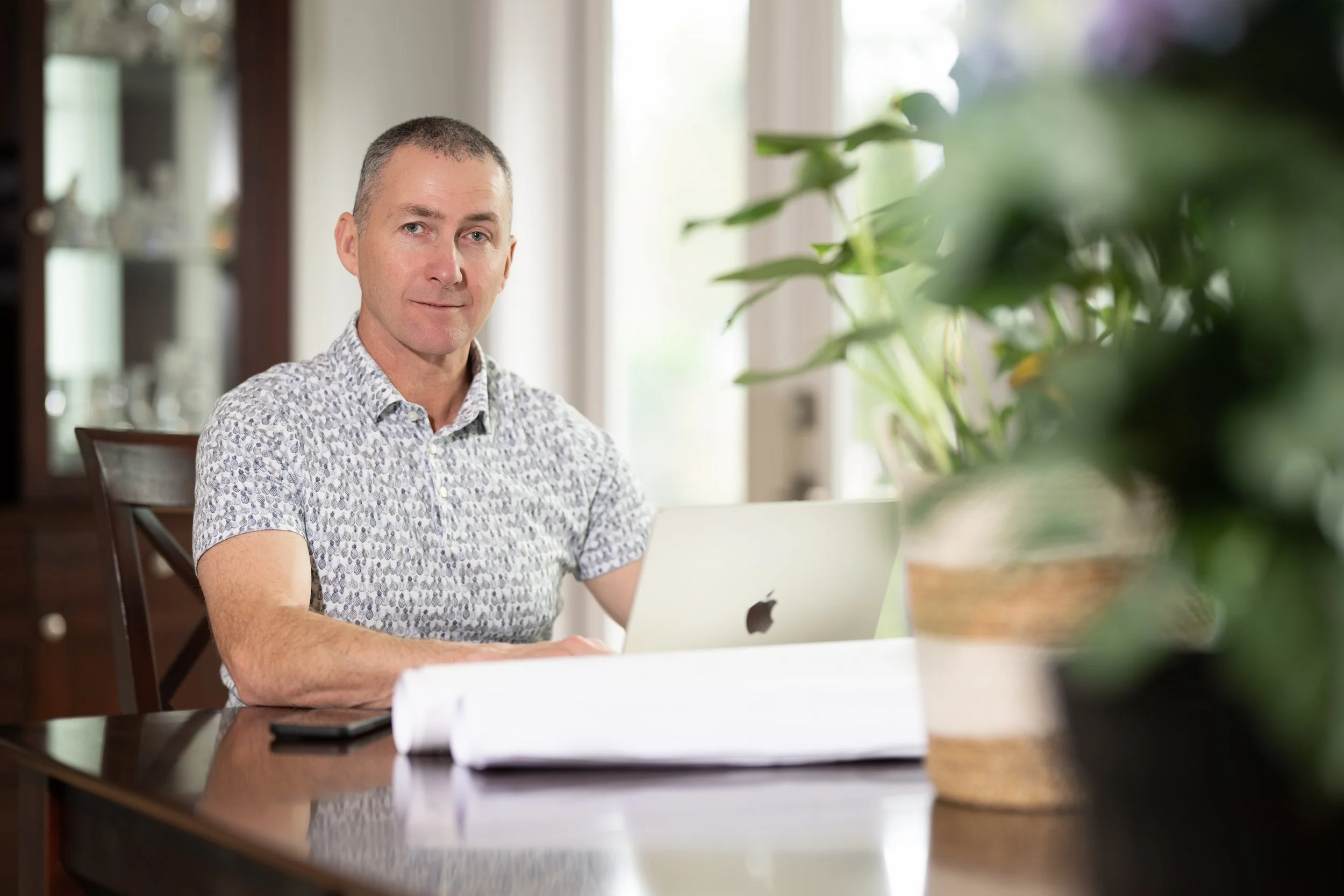 Man sitting at a wooden table with a laptop, a smartphone, and large potted plant in the foreground, in a bright room with windows.