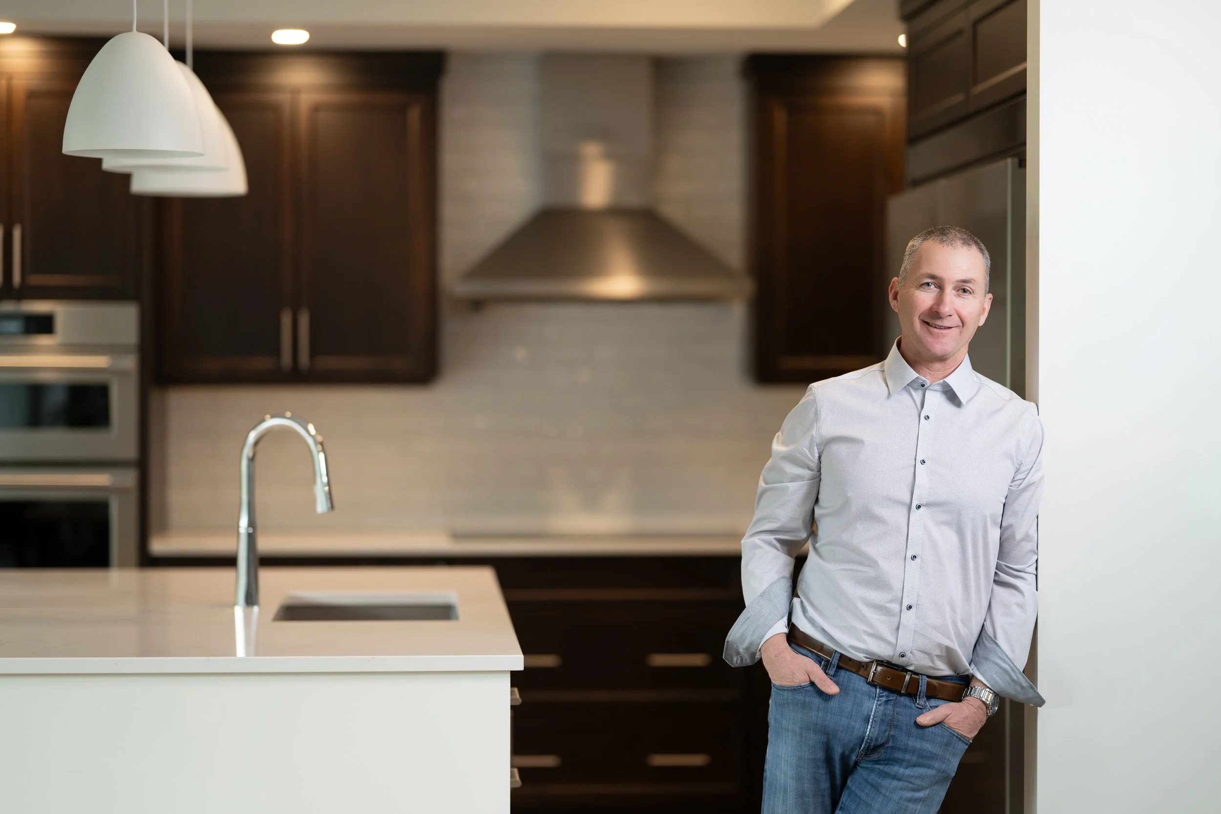 A man in a white button-up shirt and jeans leaning against a wall in a modern kitchen with dark wood cabinets and stainless steel appliances.