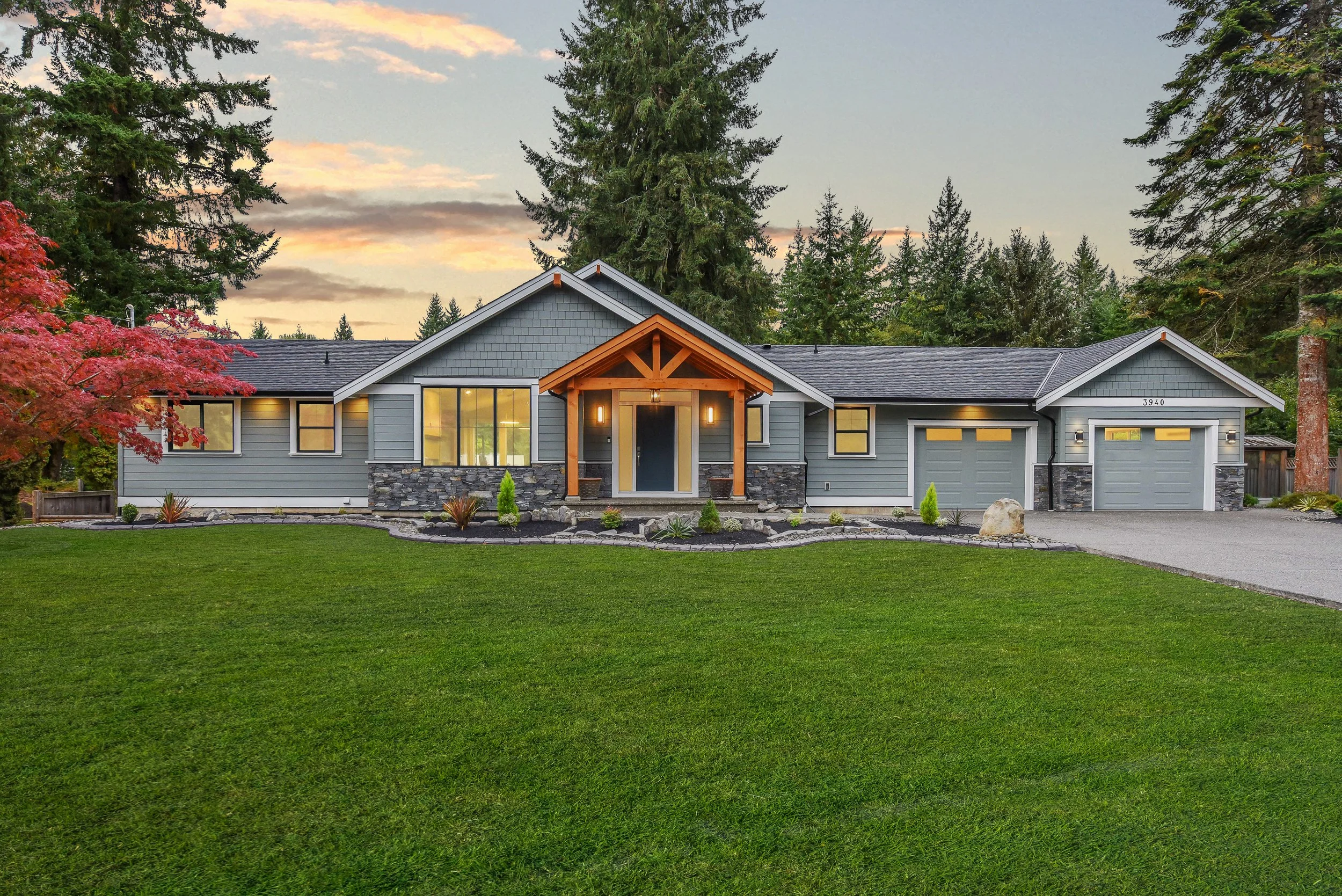 A modern single-story house with gray siding, black roof, and stone accents, surrounded by a well-maintained green lawn and trees, at sunset.