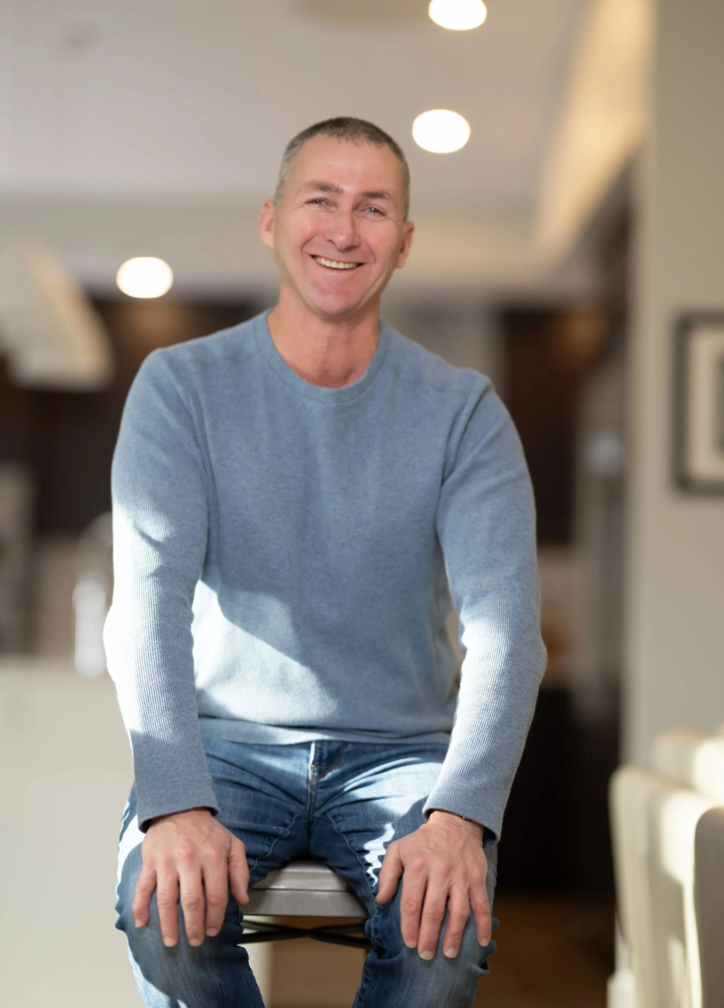 A man with short hair, smiling, wearing a light blue sweater and jeans, sitting on a chair in a cozy, well-lit room.